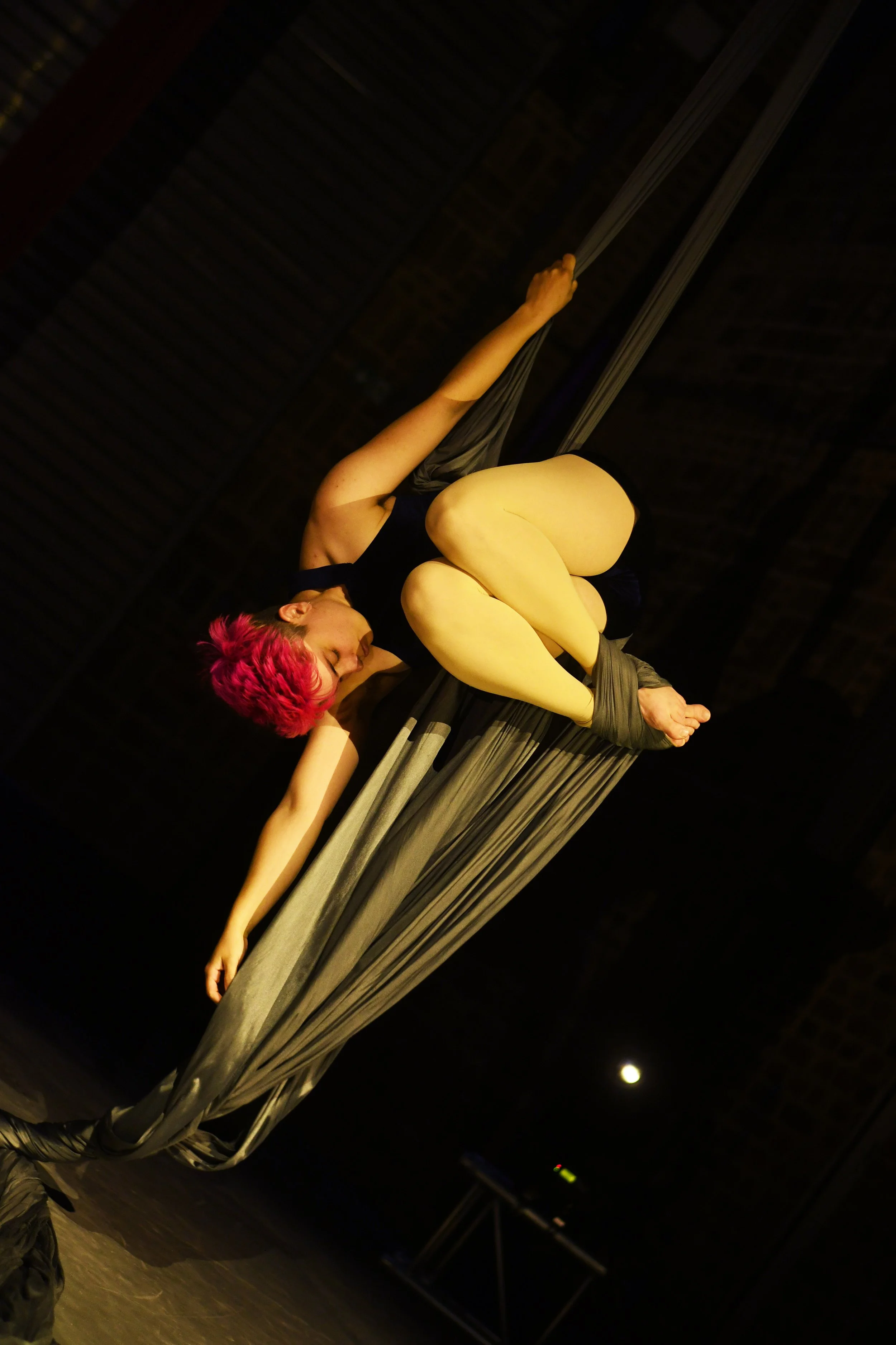 Une artiste suspendue dans un drap noir, en séance de danse aérienne ou acrobatie, avec une chevelure rose vif, dans une atmosphère sombre avec un éclairage ciblé.