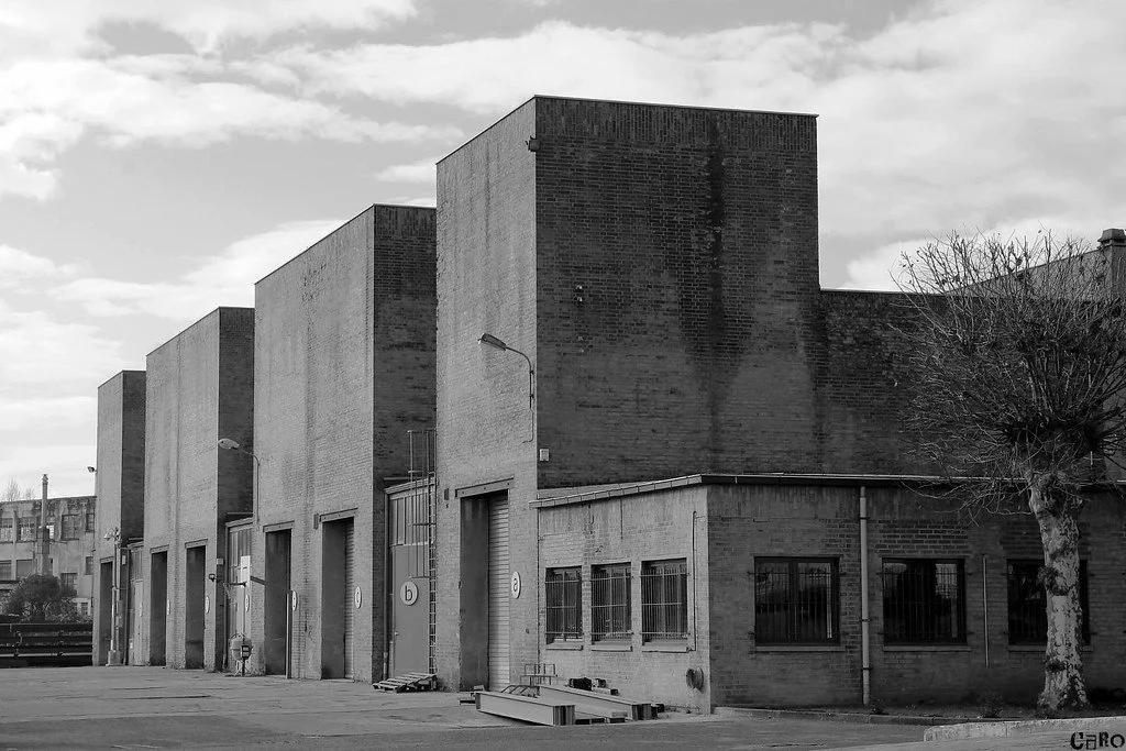 Une image en noir et blanc d'un bâtiment industriel avec plusieurs grandes portes et fenêtres, et un arbre à côté.