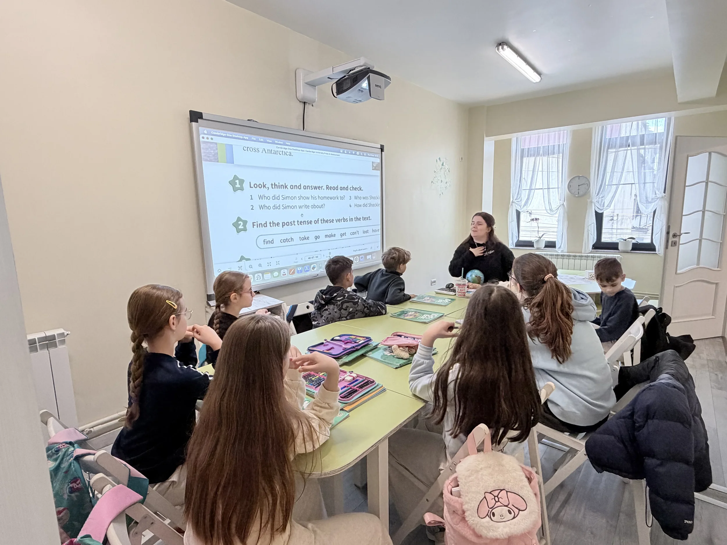 Elevi grupați la o masă lungă verde urmărind un exercițiu de gramatică proiectat pe ecranul interactiv din sala de curs pentru examenele Cambridge la York Rose Constanța