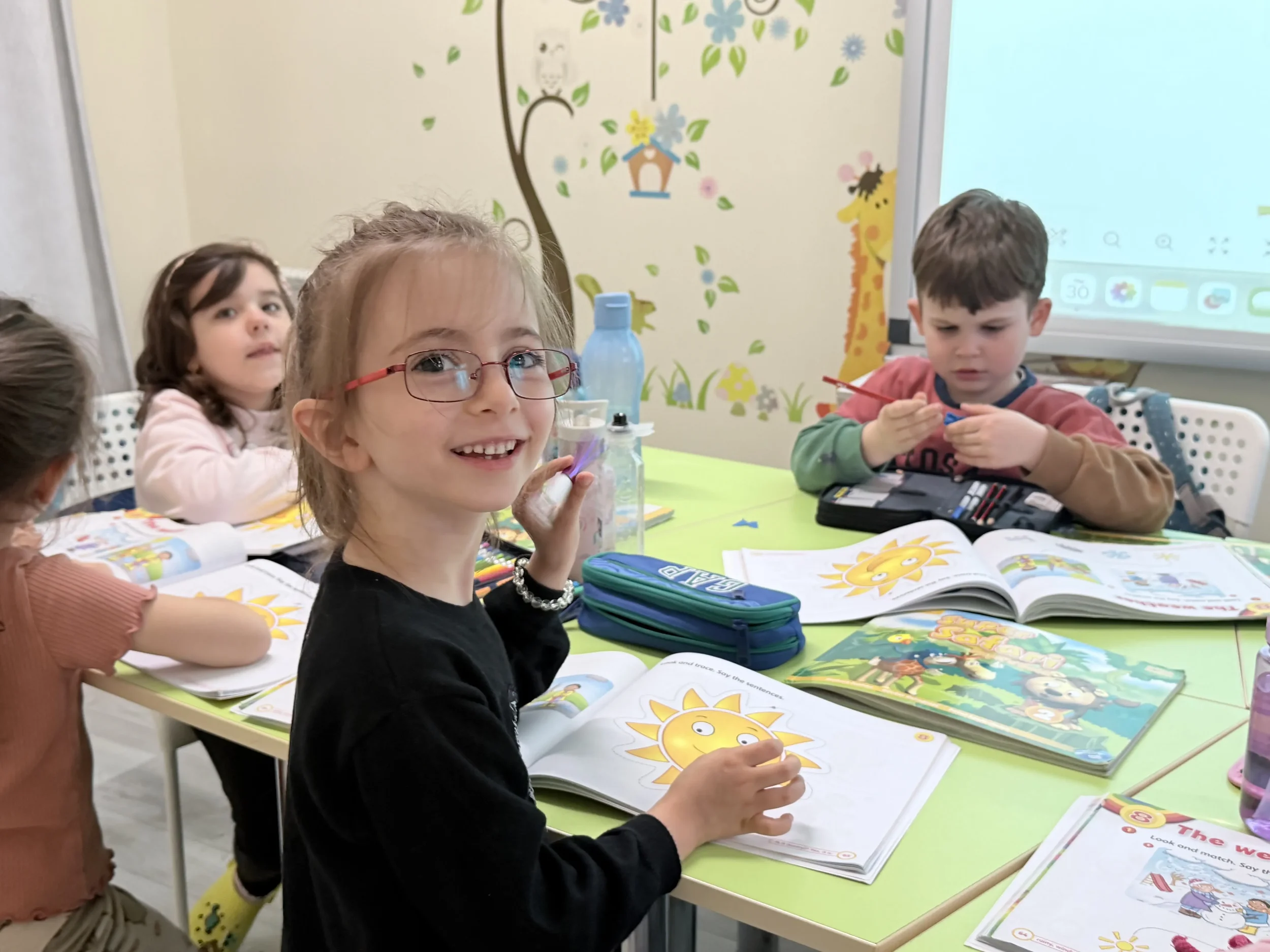 O fetiță zâmbitoare cu ochelari participă la un curs interactiv de limba engleză din manualul Super Safari, așezată la o masă verde într-o sală de clasă luminoasă.