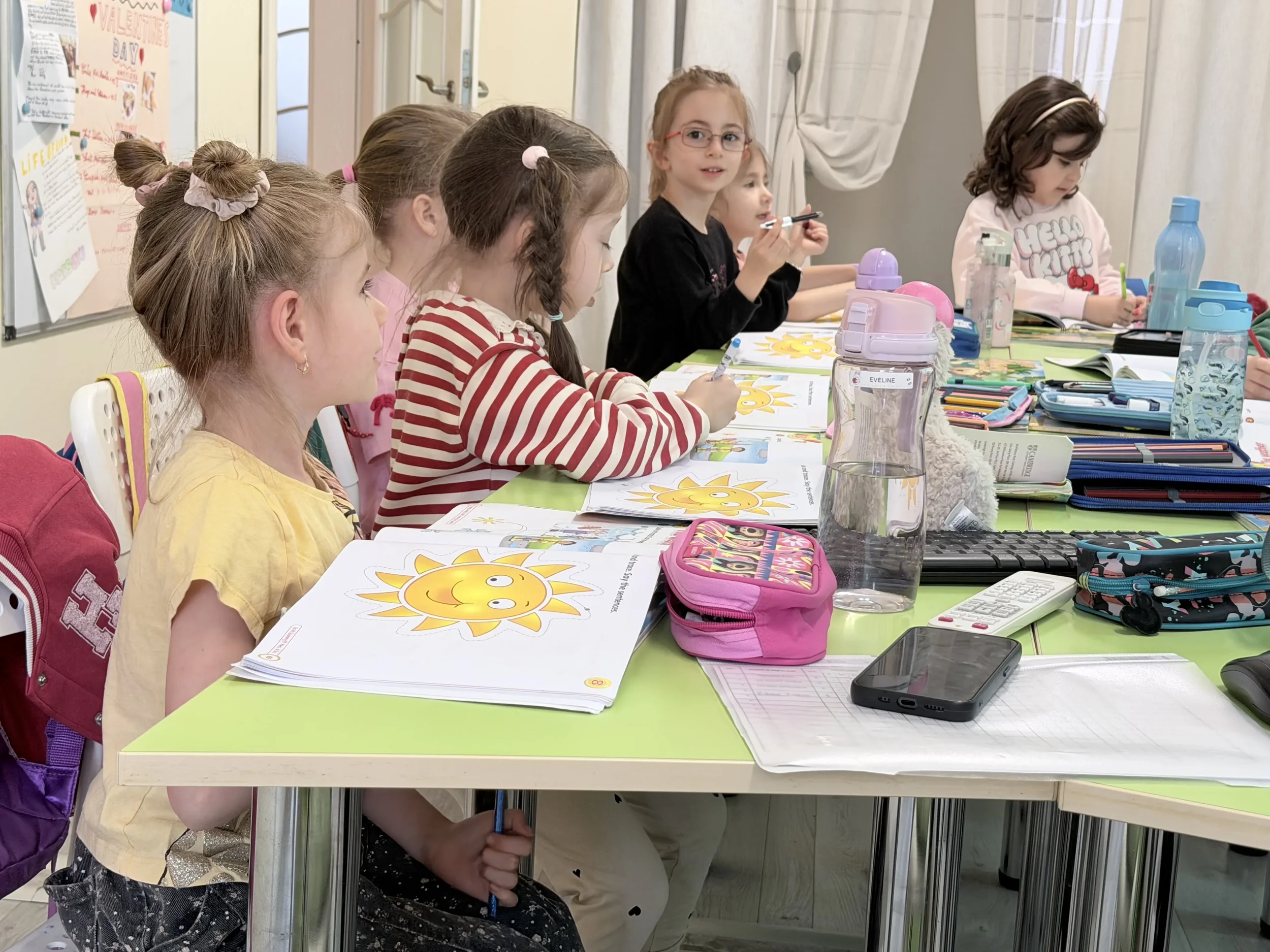 Copii de vârstă mică așezați la o masă de studiu verde, completând fișe de lucru ilustrate cu sori galbeni în cadrul unui modul de vară York Rose.