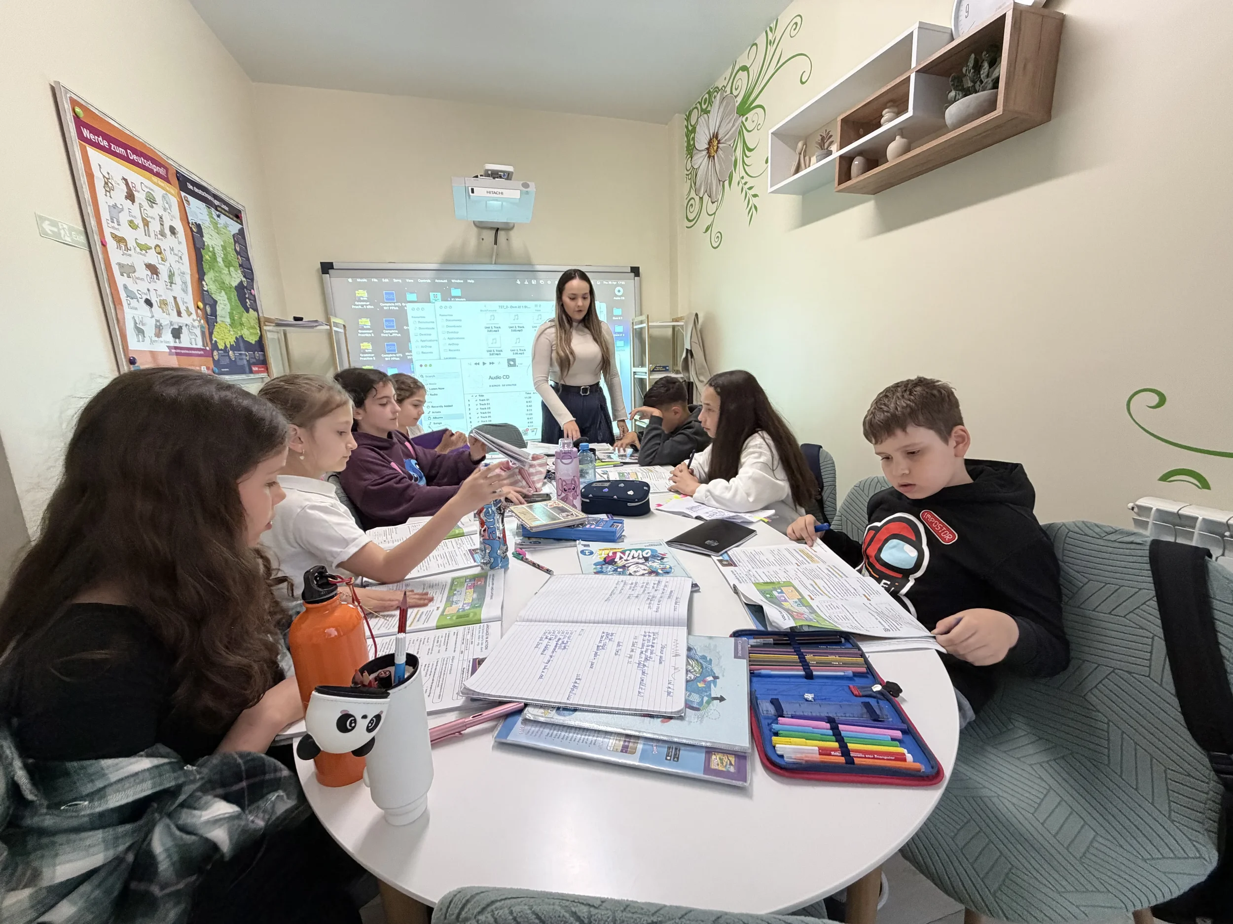 O profesoară de limba germană discută cu un grup de adolescenți în jurul unei mese albe, parcurgând exerciții pe o tablă digitală interactivă.