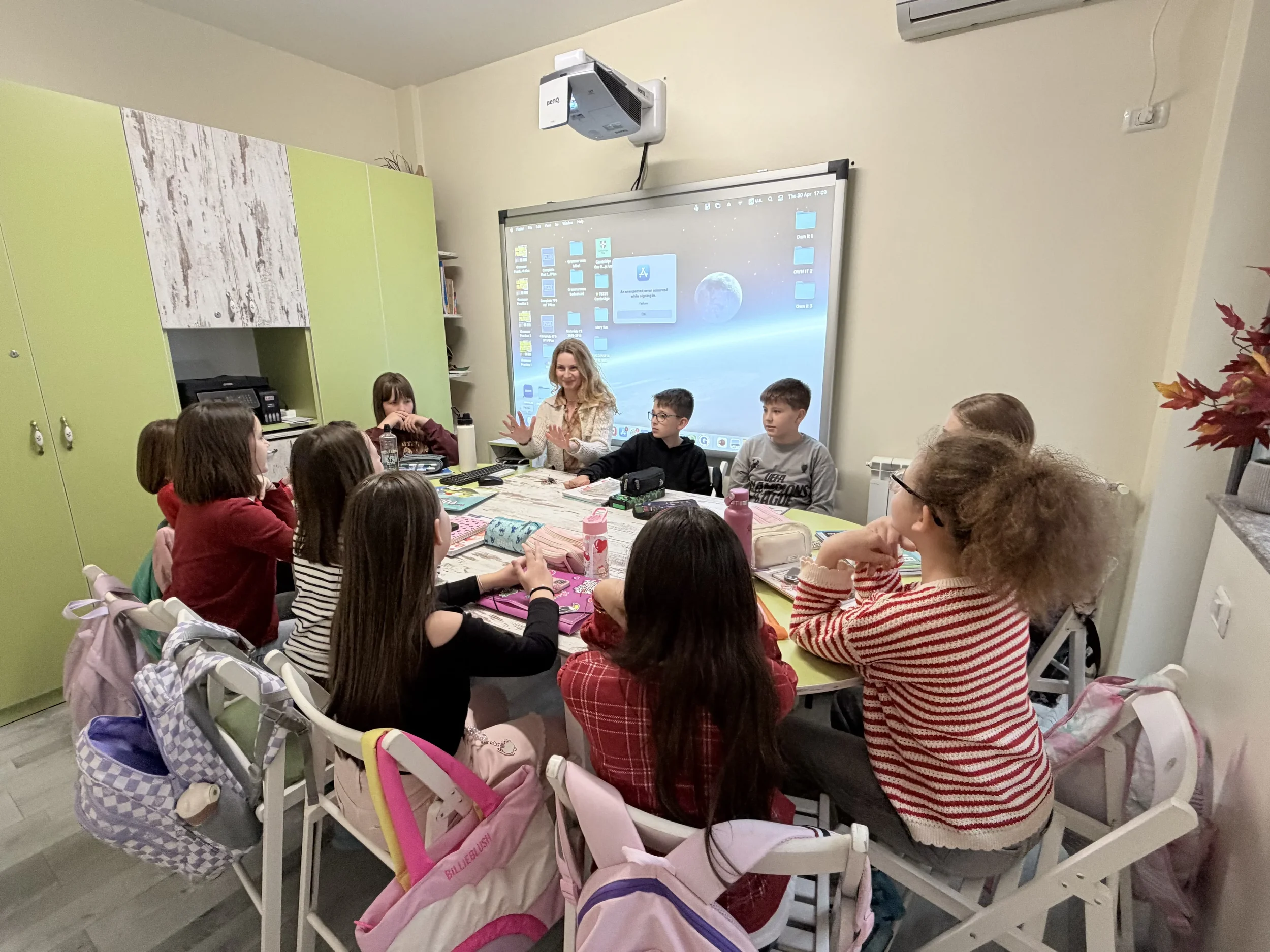Tineri cursanți așezați la o masă rotundă într-o sală de clasă modernă boutique, parcurgând un text istoric pe ecranul interactiv în cadrul pregătirii Cambridge.