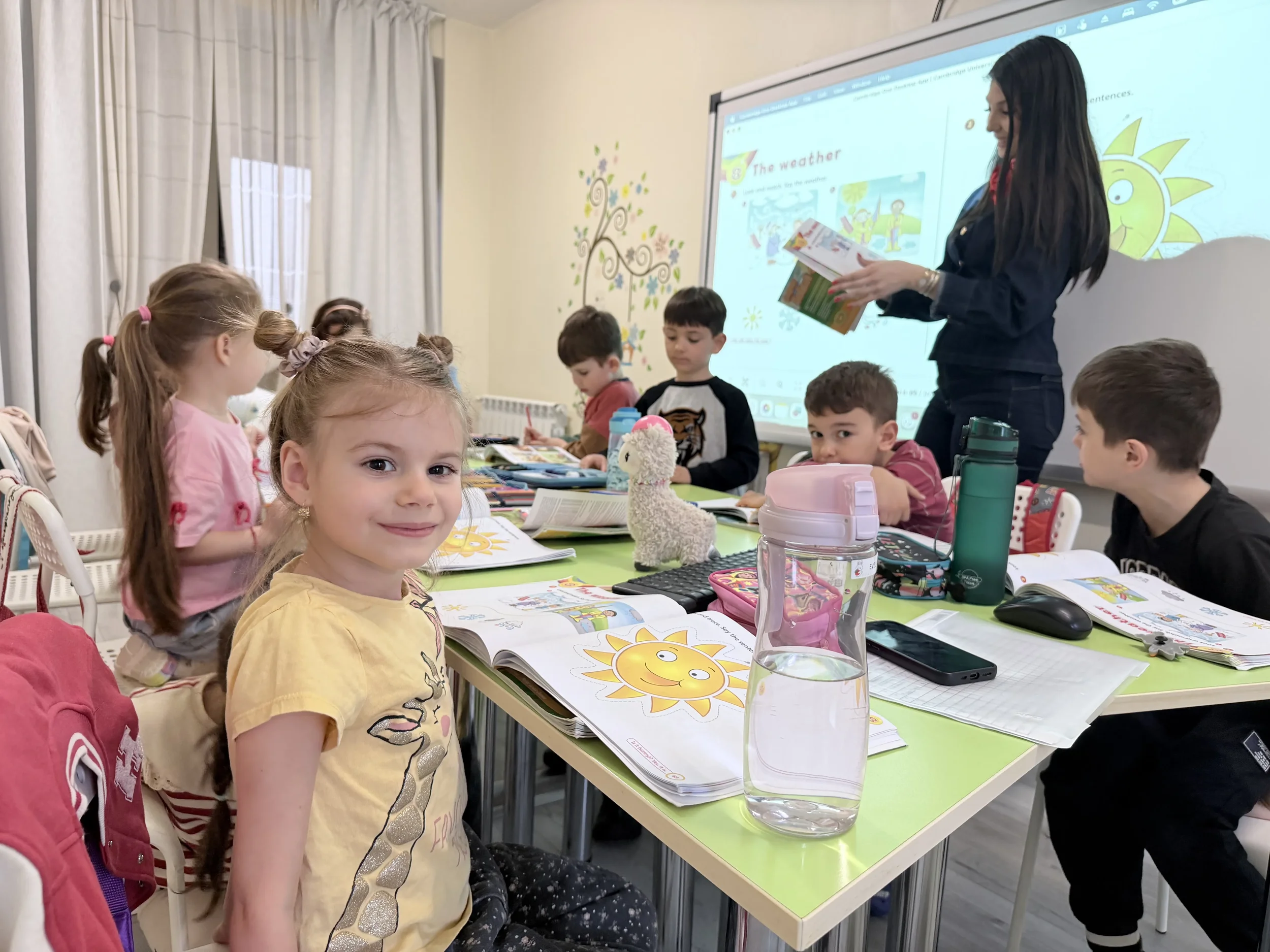 O fetiță zâmbitoare în prim-plan cu manualul deschis, în timp ce în fundal o profesoară explică lecția despre vreme la tabla interactivă.