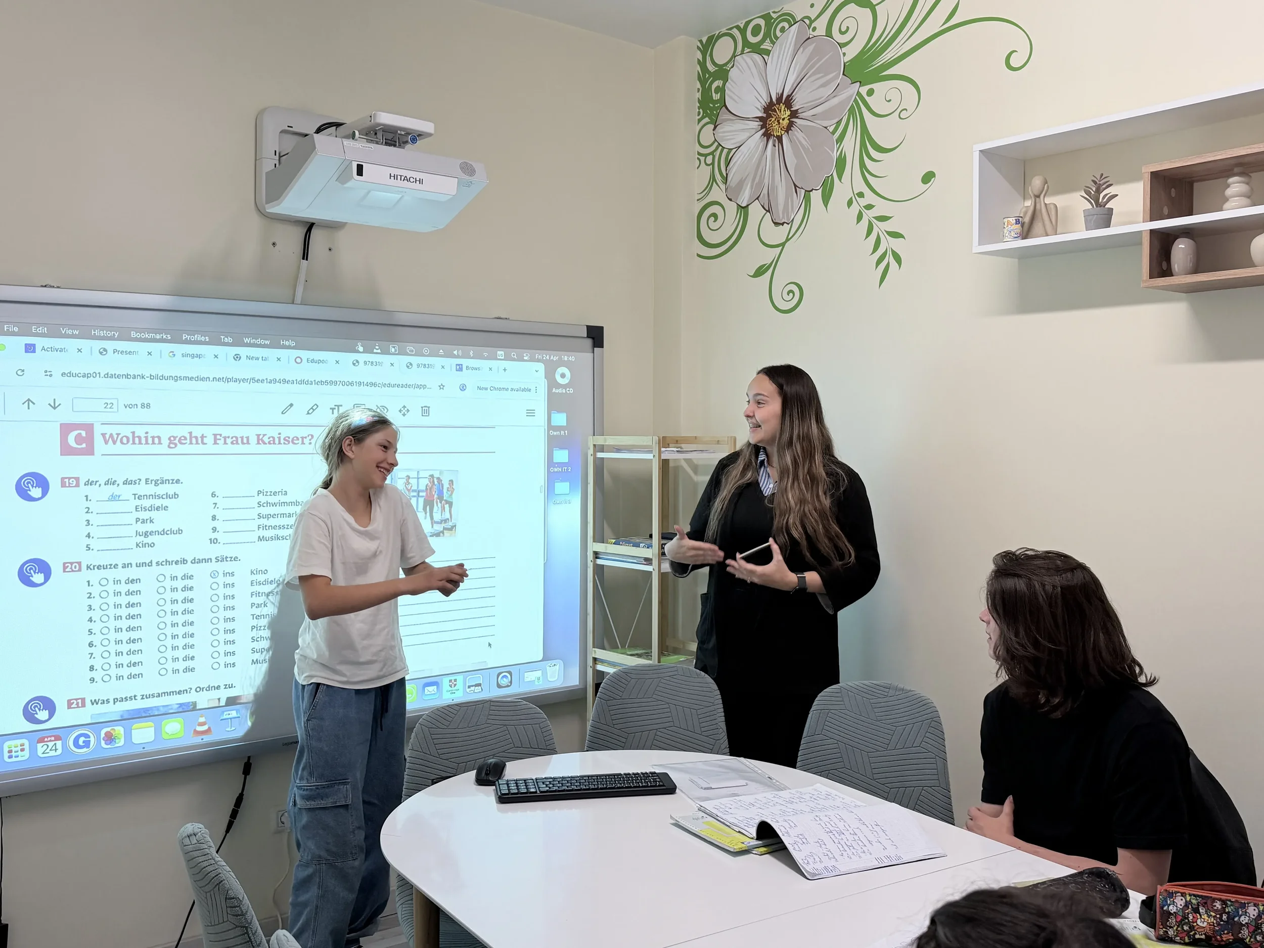 Elevă zâmbitoare la York Rose Constanța discutând cu profesoara lângă ecranul digital pe care este proiectat un exercițiu în limba germană.