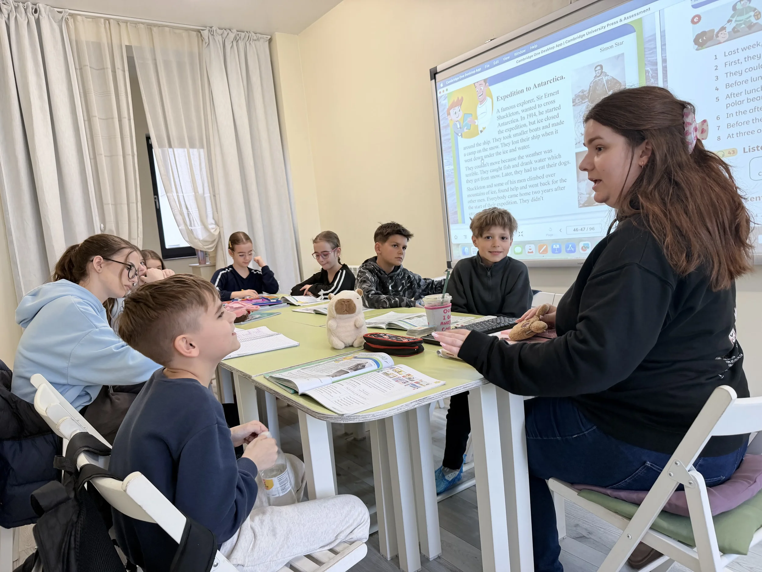 Profesoară explicând o lecție despre o expediție pe ecranul digital în fața unui grup de copii concentrați la masă pentru pregatirea Cambridge la York Rose Constanța
