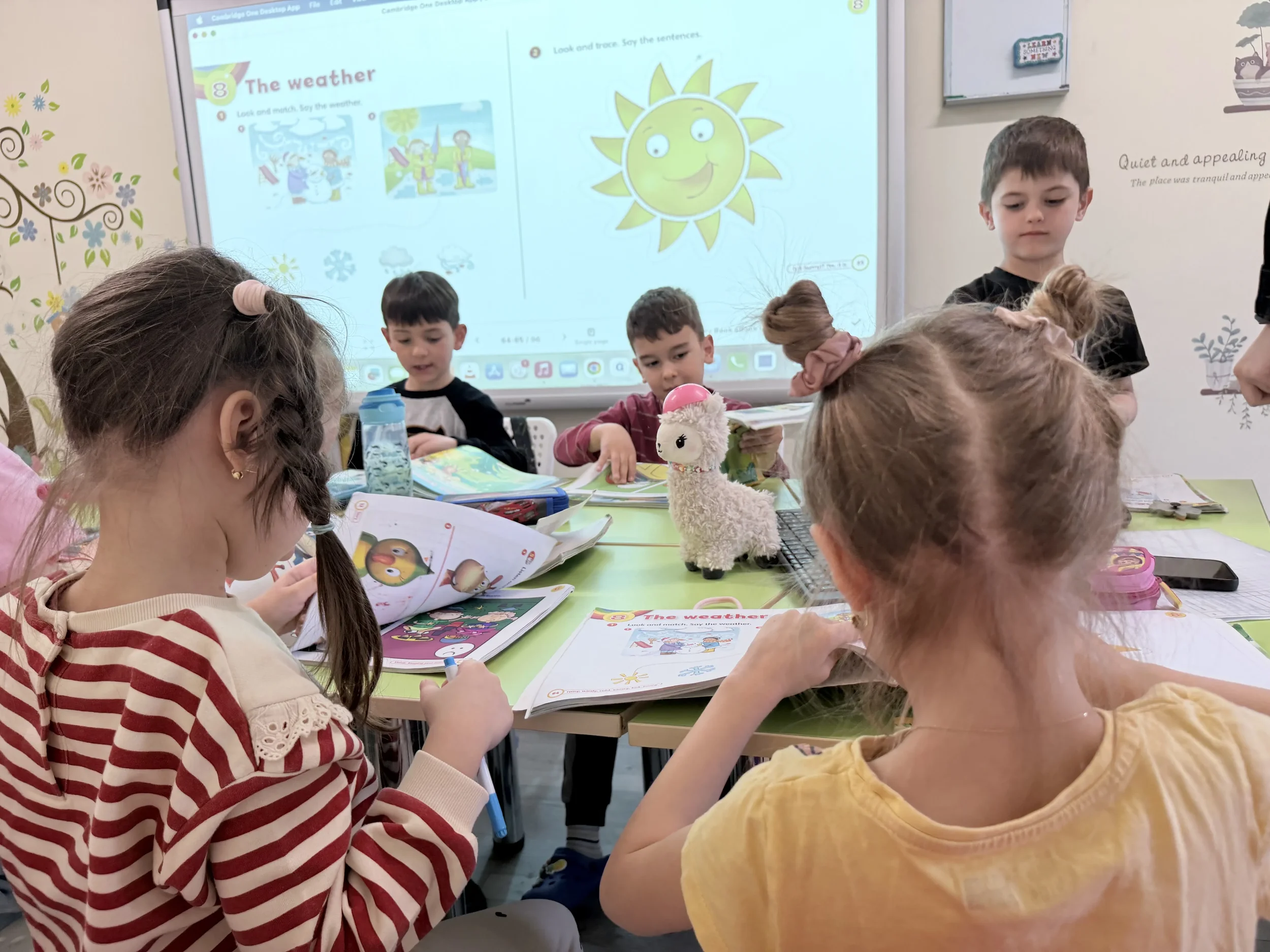 Copii care studiază din manuale ilustrate Cambridge lângă o mascotă din pluș pe masa de curs de engleza Constanta