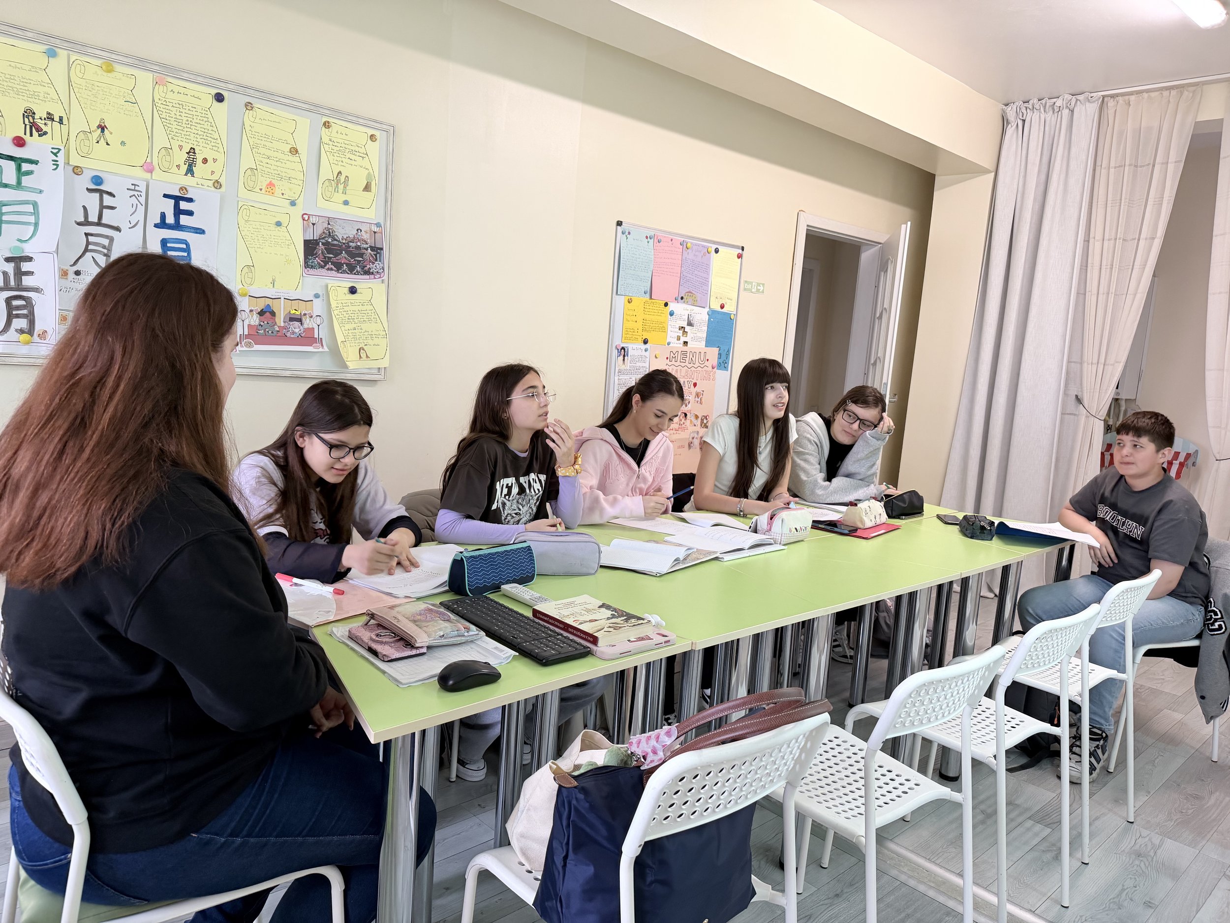 Grup de elevi adolescenți așezați la o masă lungă în timpul unui curs de limba japoneză pentru juniori în Constanța. Atmosferă caldă, de tip boutique, cu materiale didactice deschise și panou cu proiecte pe fundal.