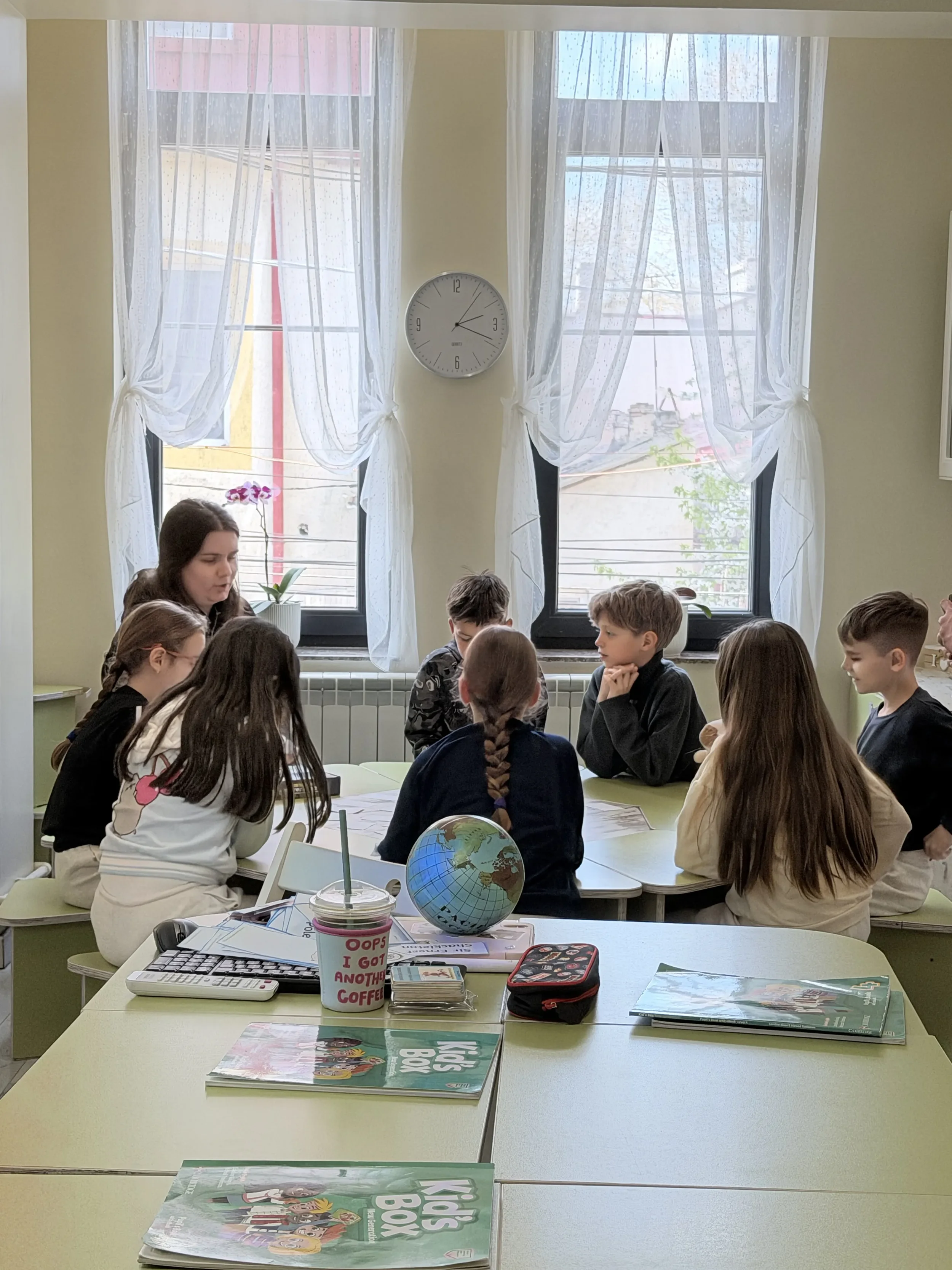 Grup de copii așezați la masa de studiu cu un glob pământesc și manuale Kids Box în fața ferestrei, ascultând profesoara la pregatirea pentru Cambridge in Constanța