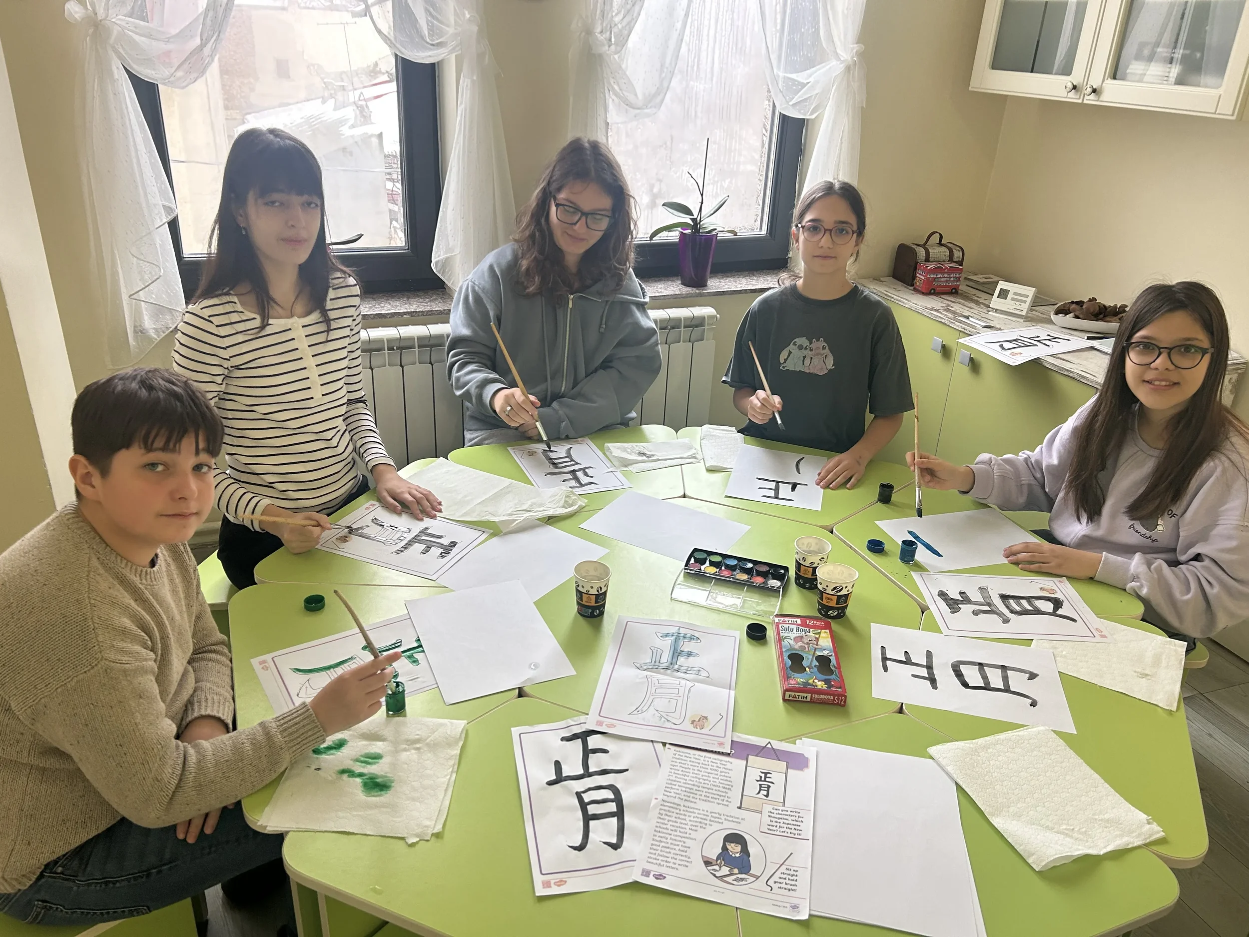 Elevi concentrați exersând scrierea caracterelor kanji cu pensule și acuarele la o masă verde în sala de curs York Rose Constanța