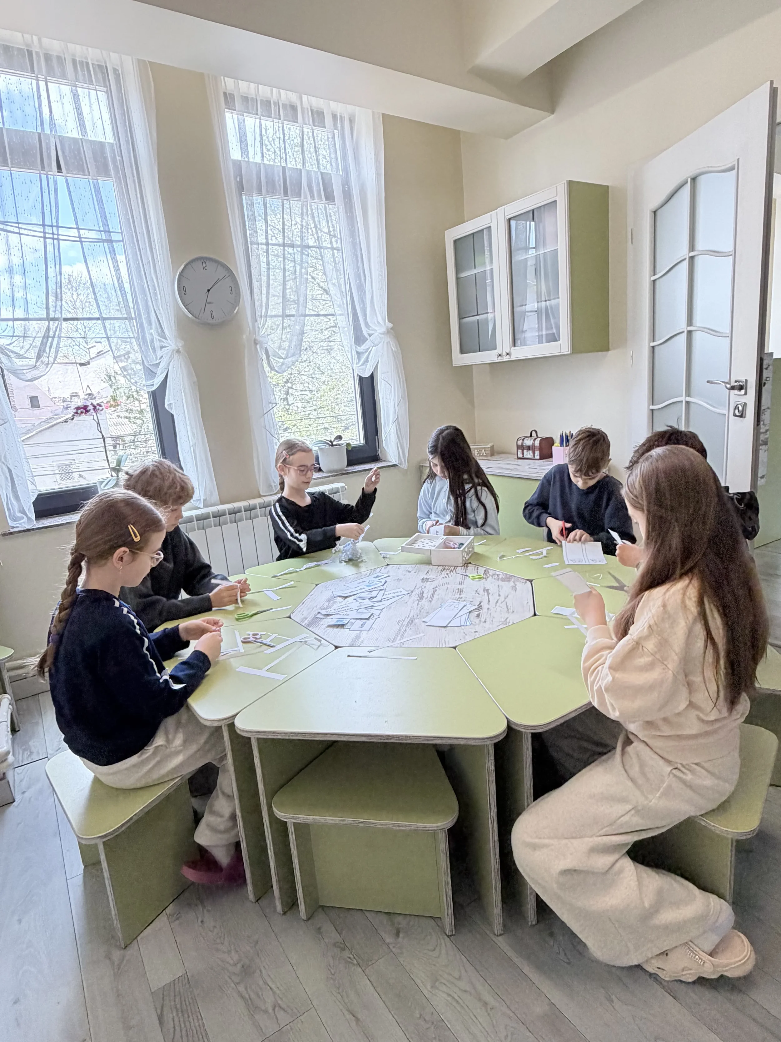 Copii așezați la o masă octogonală verde în timp ce realizează proiecte creative din hârtie la cursul de limba germană la York Rose Constanța