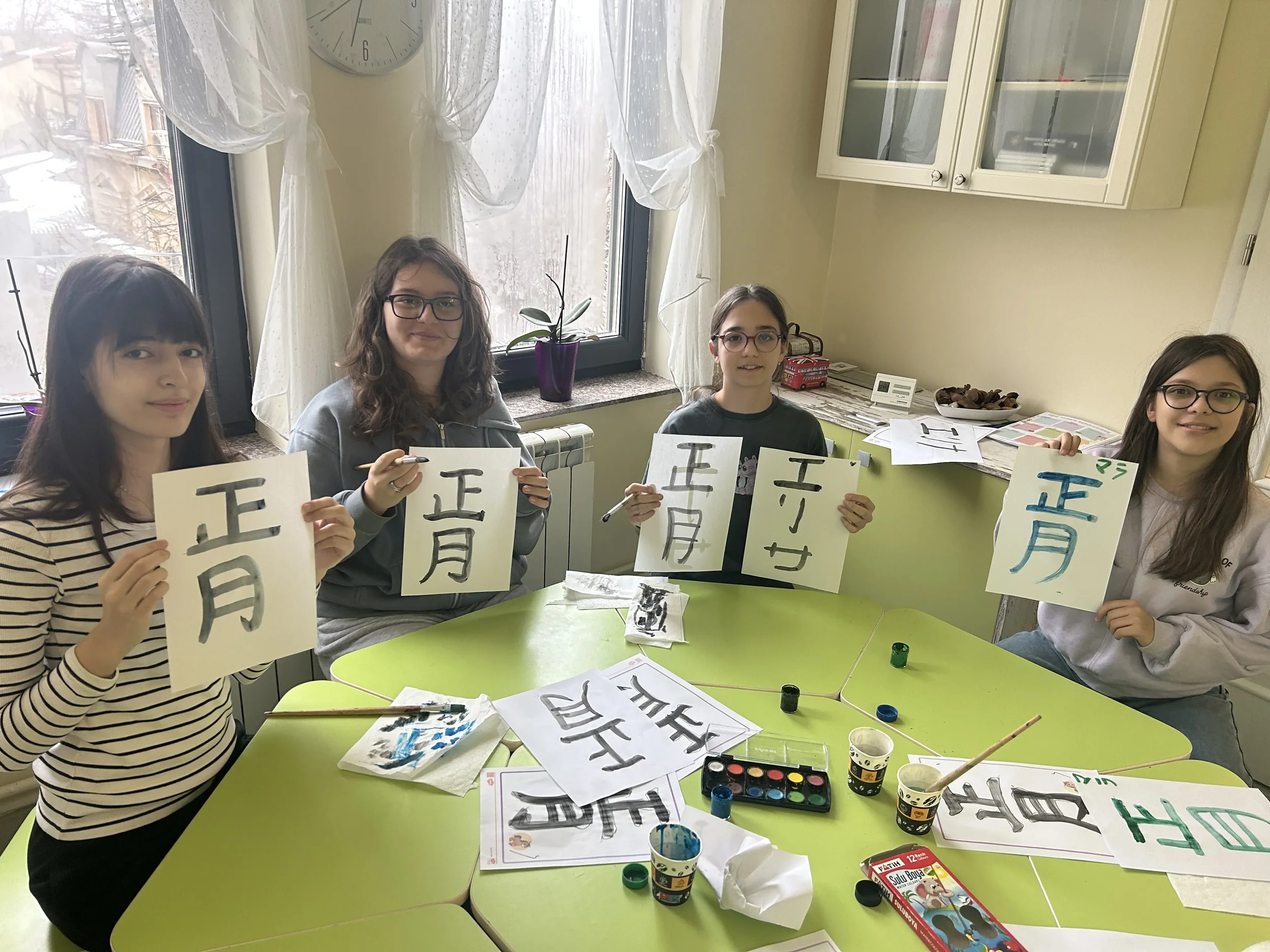 Patru eleve zâmbitoare care își prezintă lucrările de caligrafie japoneză realizate cu pensula la masa de curs la York Rose Constanța