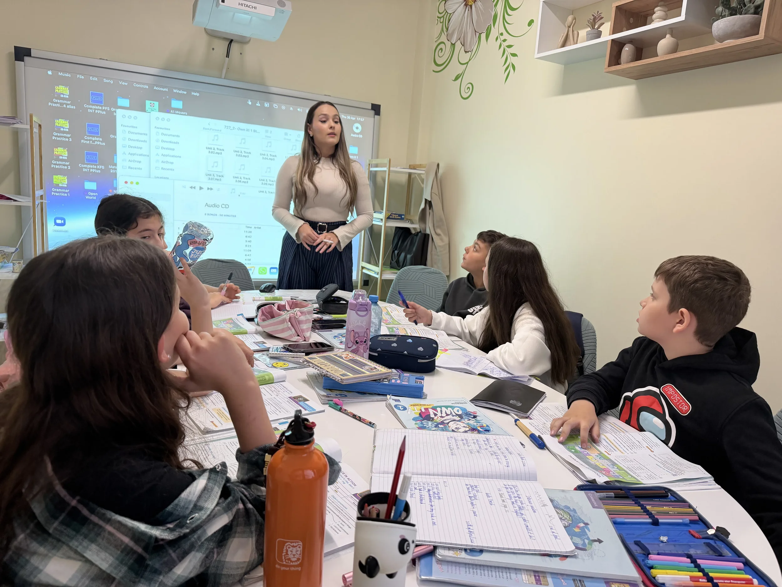 Profesoară în picioare la pregǎtirea pentru Cambridge, coordonând o lecție dinamică pentru elevii așezați la masa de curs cu manuale deschise și penare