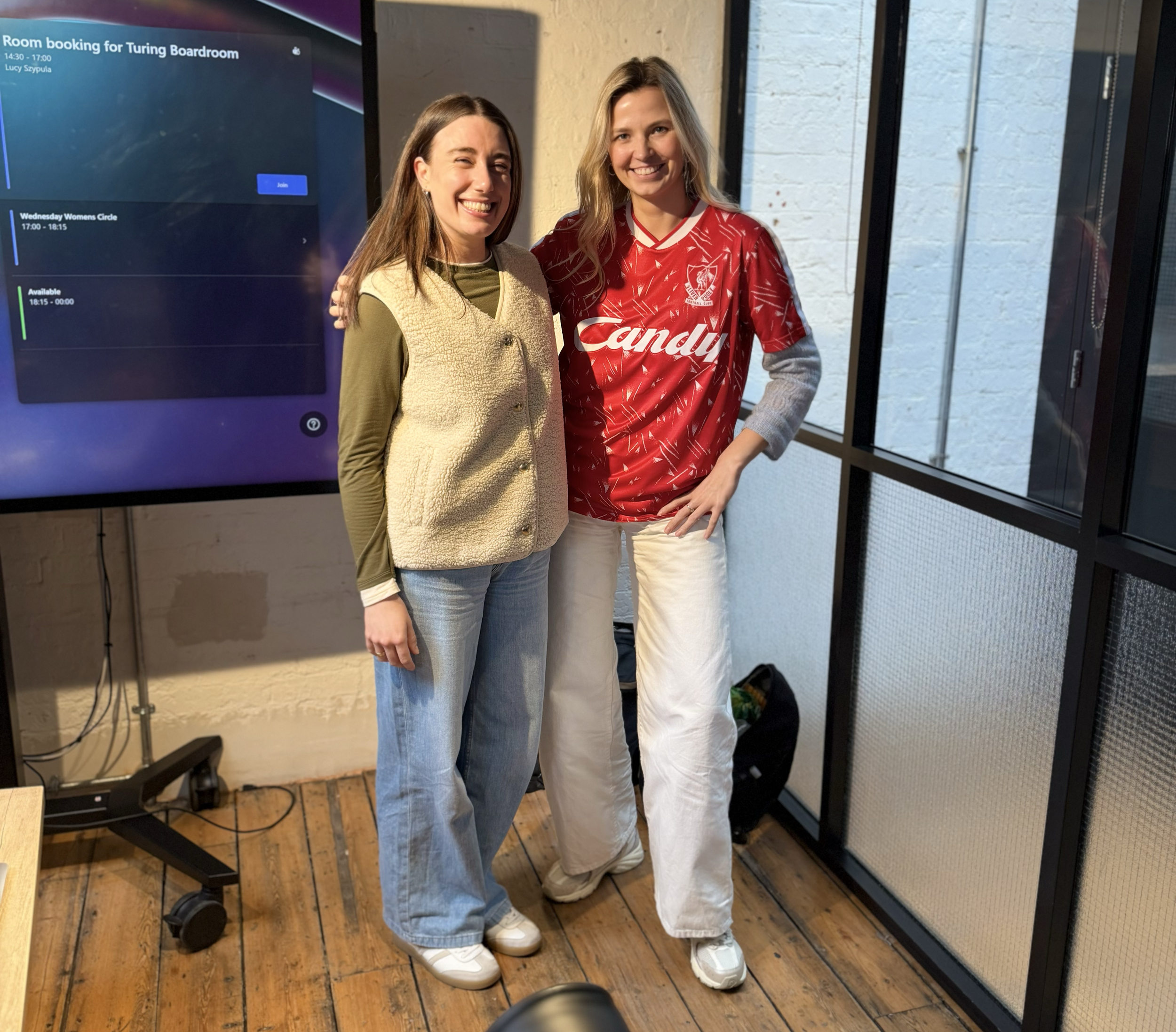 Two young women smiling and standing close together indoors near a glass door and a large screen displaying room booking information. One wears a beige vest over an olive green shirt, the other wears a red sports jersey with the word 'Candy' and a logo.