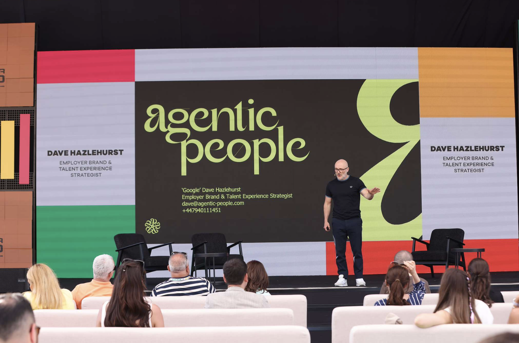 A speaker standing on stage at a conference or event, in front of a large screen displaying the words 'agentic people' and information about Dave Hazlehurst, an employer brand and talent experience strategist. There are seated audience members watching the presentation.