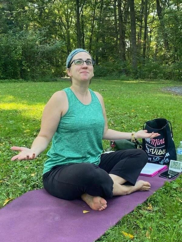 A woman sitting cross-legged on a purple yoga mat outdoors in a grassy area with trees in the background. She is wearing glasses, a blue headband, a sleeveless green top, and black pants. She appears to be meditating or practicing mindfulness, with a calm expression and her arms relaxed at her sides. There are items nearby including a black bag with writing, a pink notebook, a smartphone, and a small electronic device.