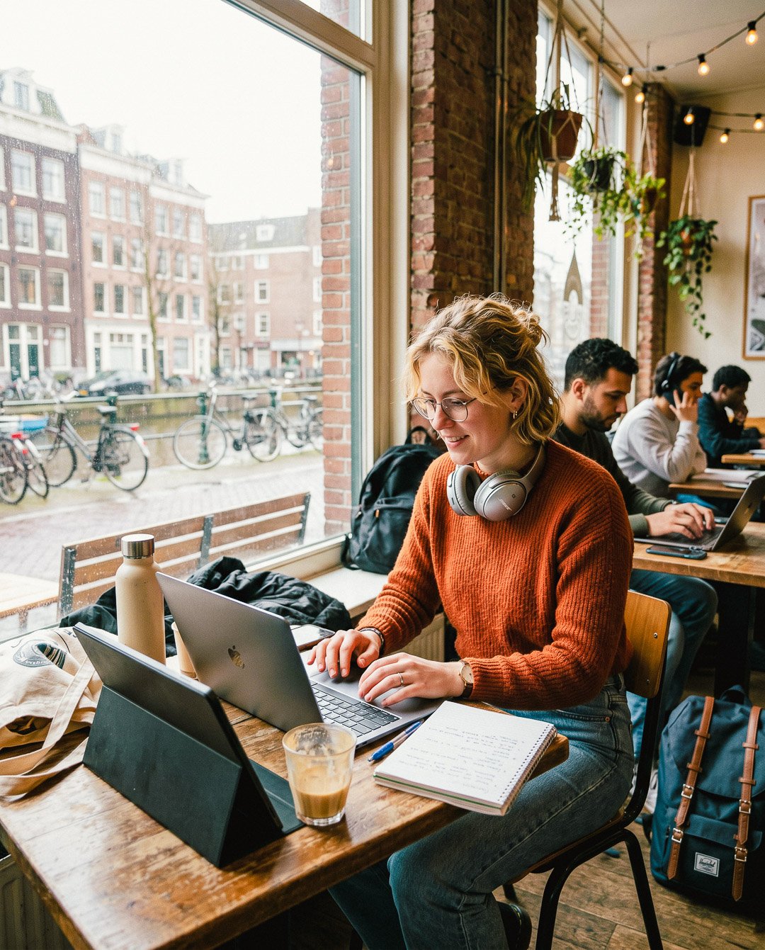 Vrouw met blond haar en glazen werkt met laptop in een café, andere mensen werken en luisteren naar muziek op achtergrond, grote ramen met uitzicht op een straat met fietsen.
