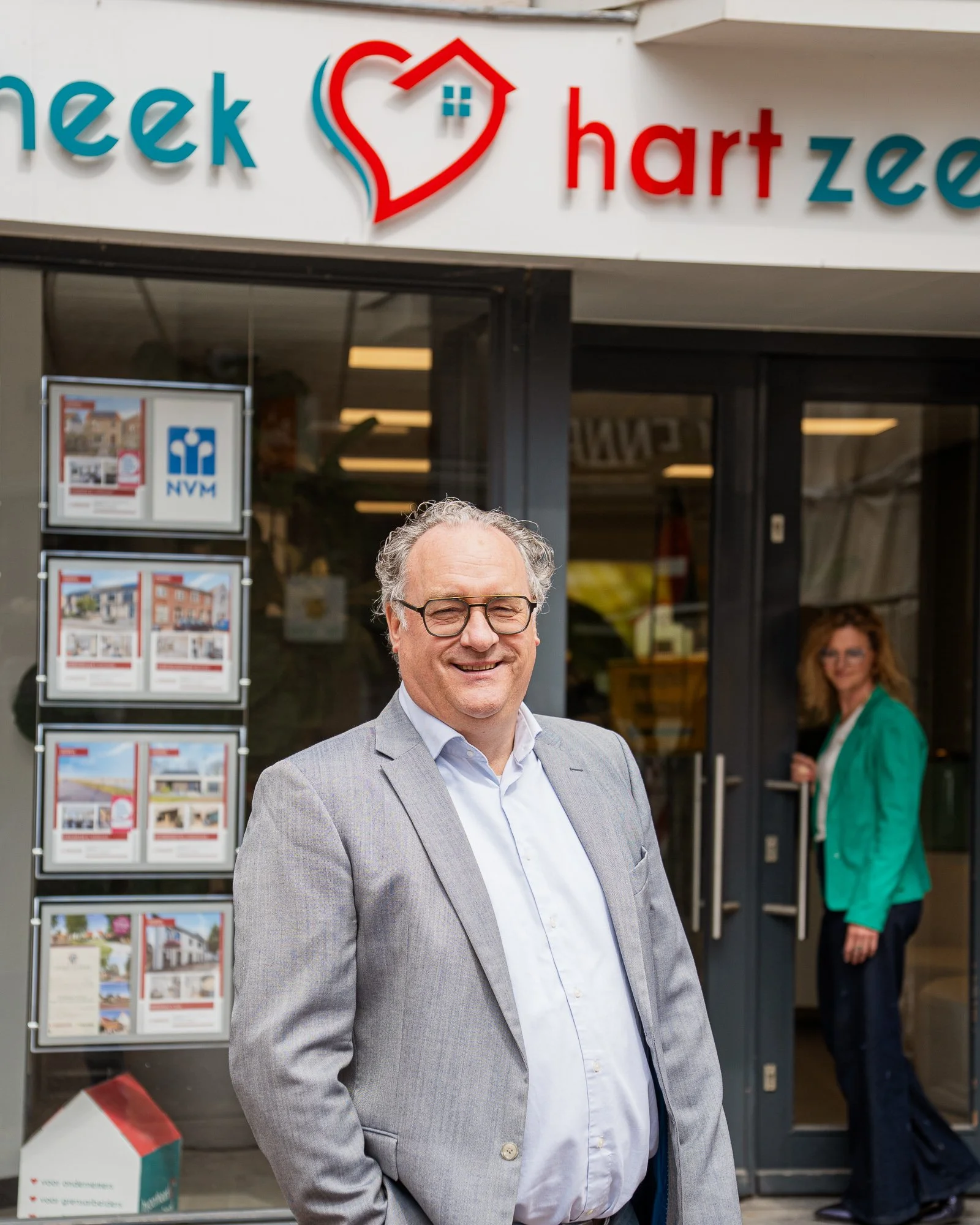 Man in grijs pak staat voor het gebouw 'Huis & Hart' met een vrouw in groene jas in de achtergrond.