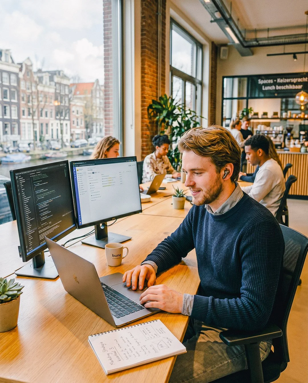 Man werkt aan laptop in een modern kantoor met collega’s op de achtergrond, grote ramen, groene planten, en uitzicht op historische gebouwen.
