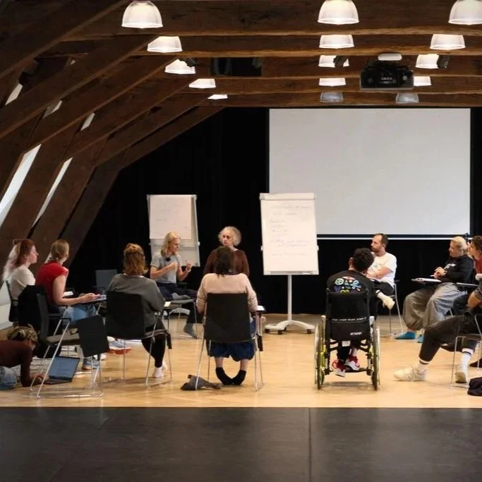 Eine Gruppe von Menschen sitzt im Kreis in einem Raum mit Holzdecke, an zwei Whiteboards steht ein Gespräch. Es sieht nach einer Gruppen- oder Teambesprechung aus.