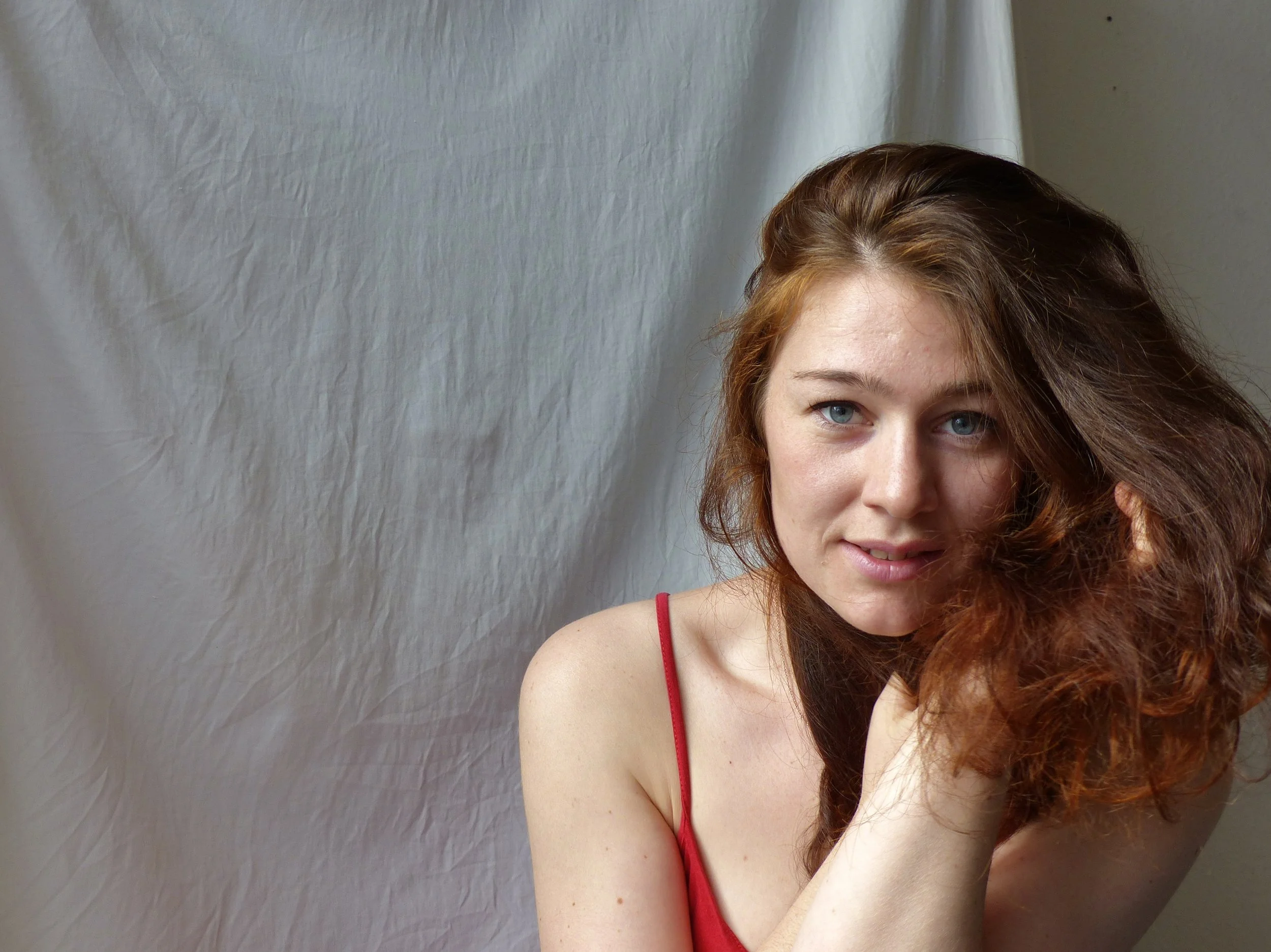 Eine junge Frau mit roten, lockigen Haaren und blauen Augen trägt ein rotes Top. Sie sitzt vor einem weißen Hintergrund und schaut in die Kamera.