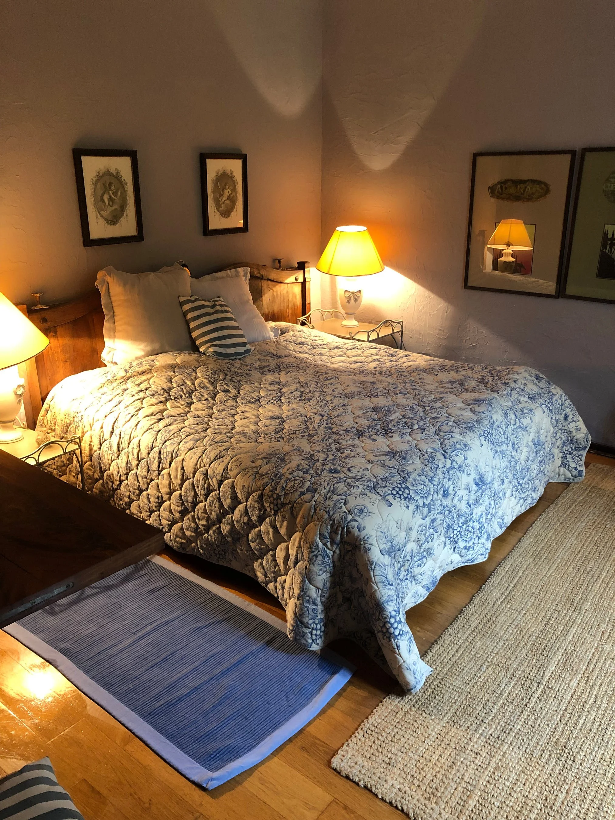 Ein gemütliches Schlafzimmer mit einem großen Bett, das mit einem Blumenmuster-Quilt bedeckt ist. Zwei Nachttische mit Lampen flankieren das Bett. An den weißen Wänden hängen drei Bilderrahmen. Der Boden ist aus Holz, mit einem kleinen blauen Teppich