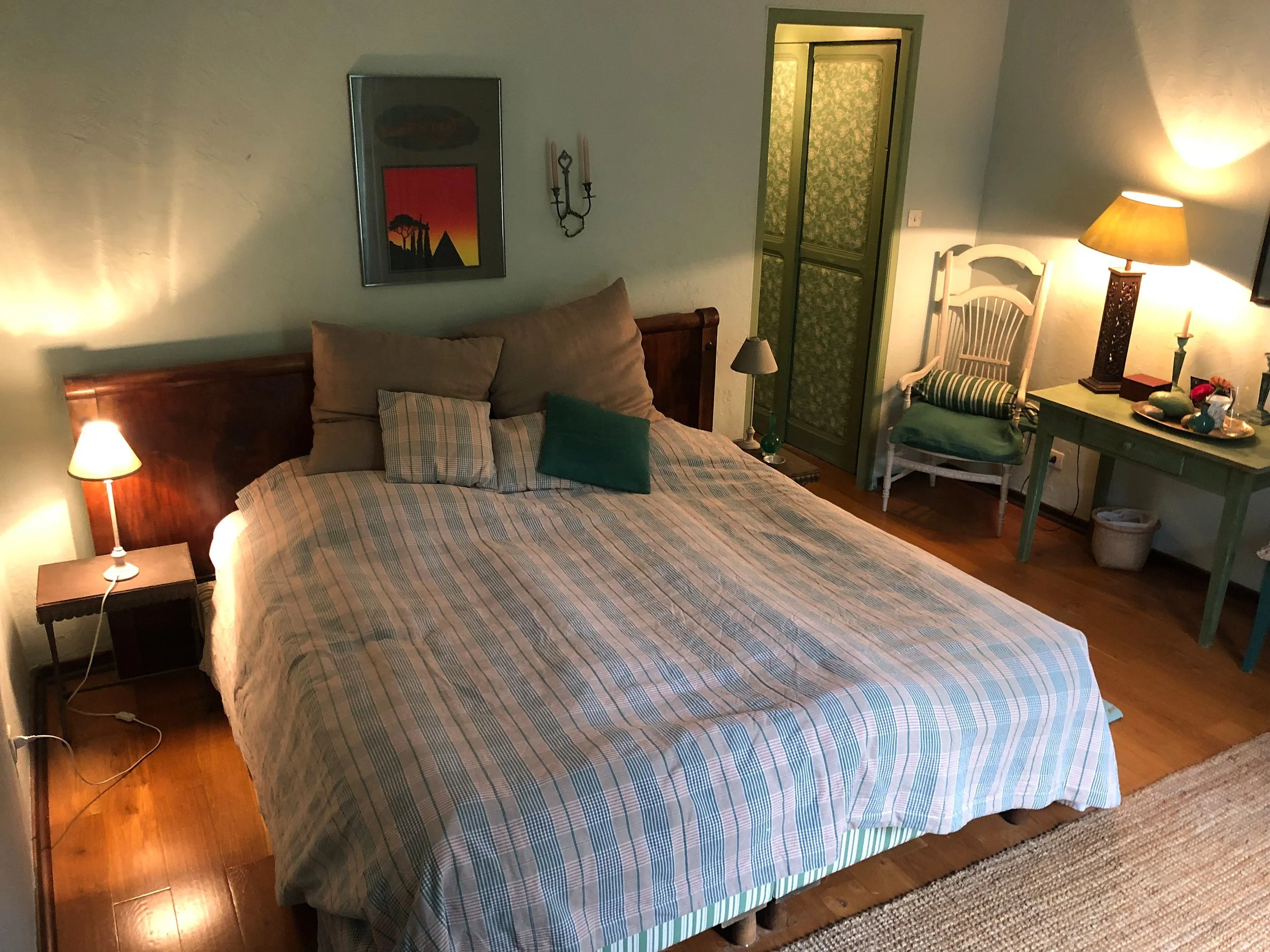 Gemütliches Schlafzimmer mit Doppelbett, Nachttischen mit Lampen, einem Sessel mit Kissen, einem grünen Beistelltisch mit Lampe, einer Schranktür und Dekorationen an den Wänden.