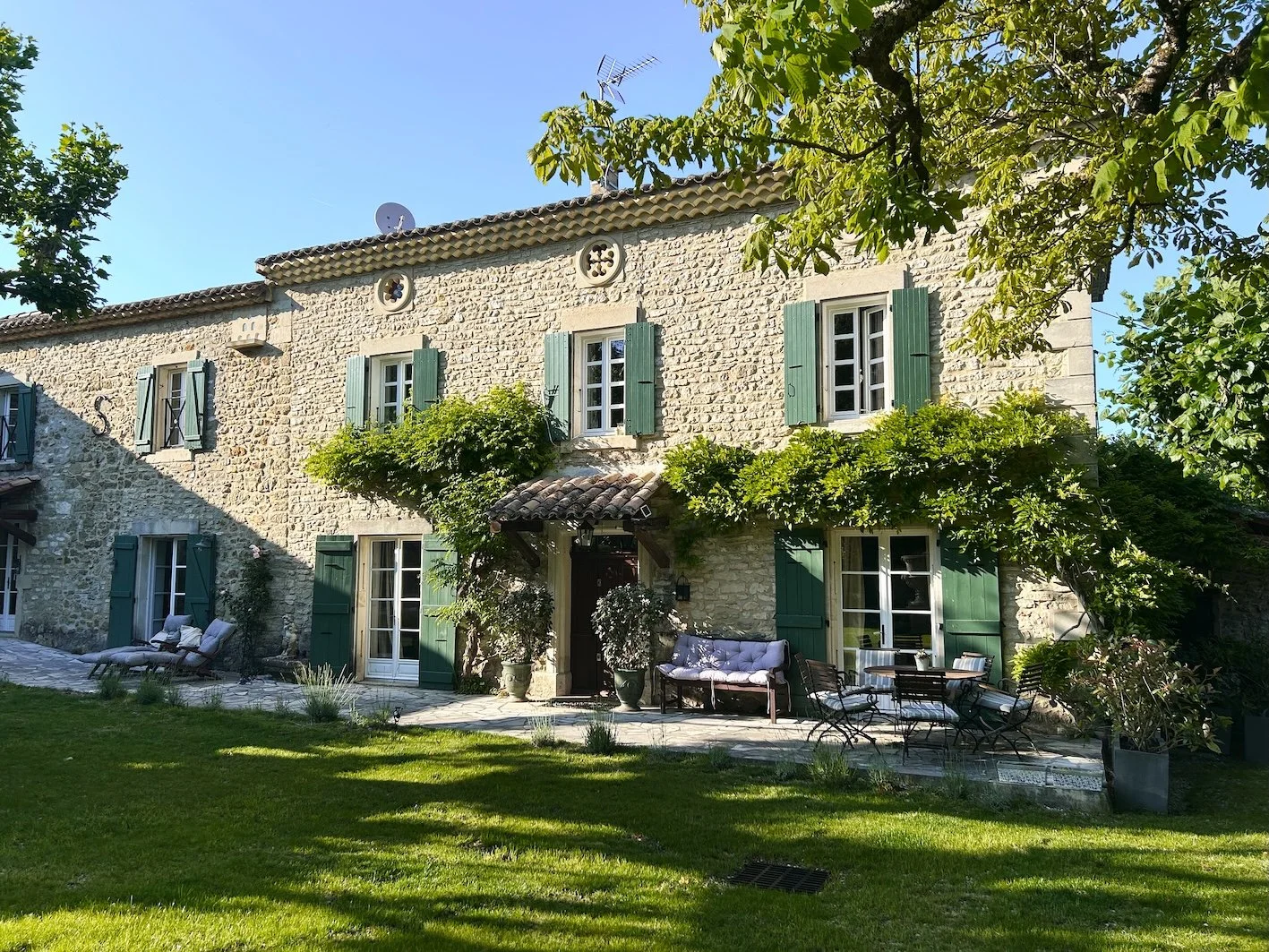 Ein charmantes Haus mit Steinmauern, grünen Fensterläden und einer gemütlichen Terrasse im Garten, umgeben von Bäumen und Pflanzen bei sonnigem Wetter.