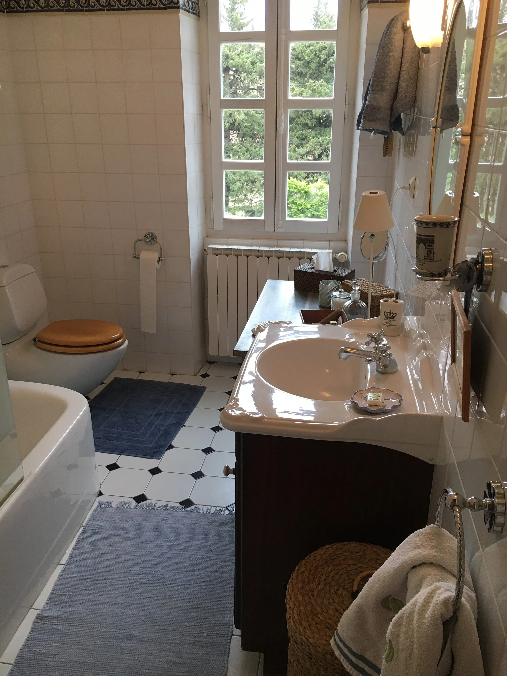 Ein Badezimmer mit einer weißen Badewanne, einem weißen Spiegelschrank, einem Waschbecken, einer Toilette mit braunem Sitz, einem Fenster mit Blick auf Bäume, einem grauen Teppich vor der Badewanne, einem kleinen Tisch mit Dekorationen und Handtücher
