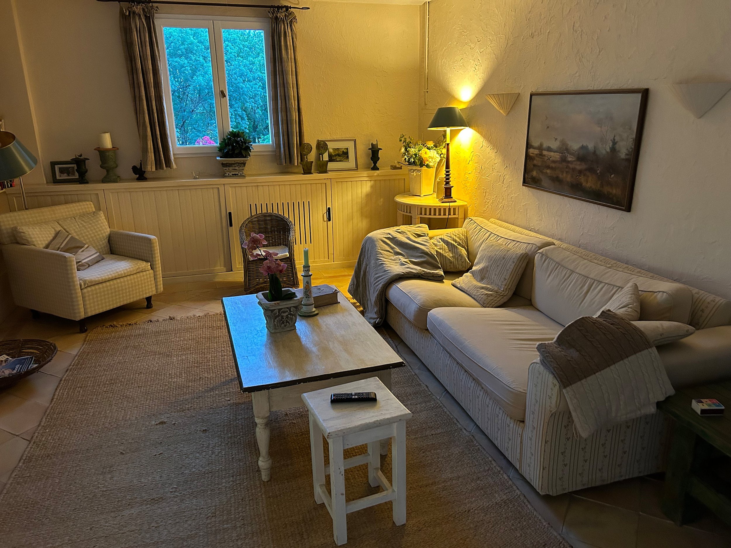 Gemütliches Wohnzimmer mit einer großen weißen Couch, einem weißen Sessel, einem Eichenholztisch, einem kleinen weißen Beistelltisch, Wandlampen, einem Gemälde und einer Fensteransicht auf Bäume bei Dämmerung.