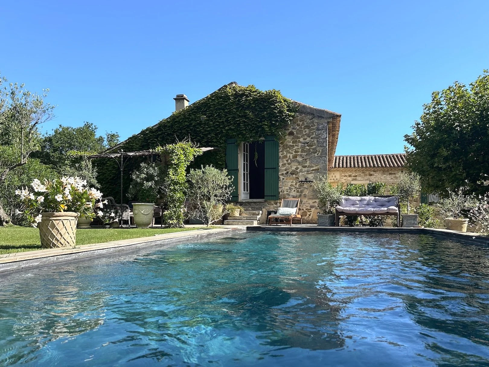 Ein Pool im Garten vor einem Steinhaus, umgeben von Pflanzen und Gartenmöbeln, bei Sonnenschein unter einem blauen Himmel.