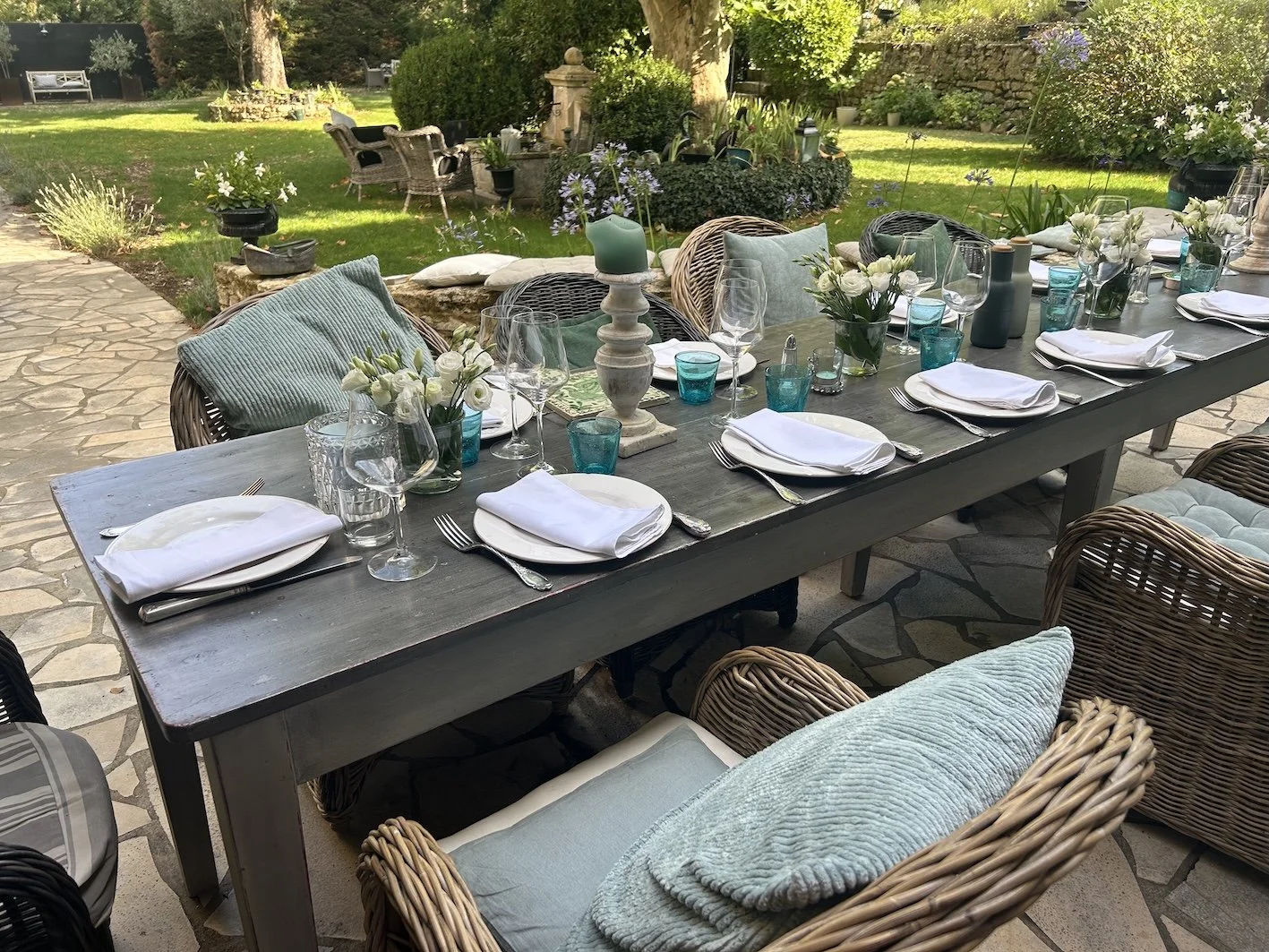 Ein festlich gedeckter Tisch im Garten mit weißen Tellern, Gläsern und Blumenarrangements, umgeben von Stühlen mit Kissen, im Hintergund Bäume und Pflanzen.