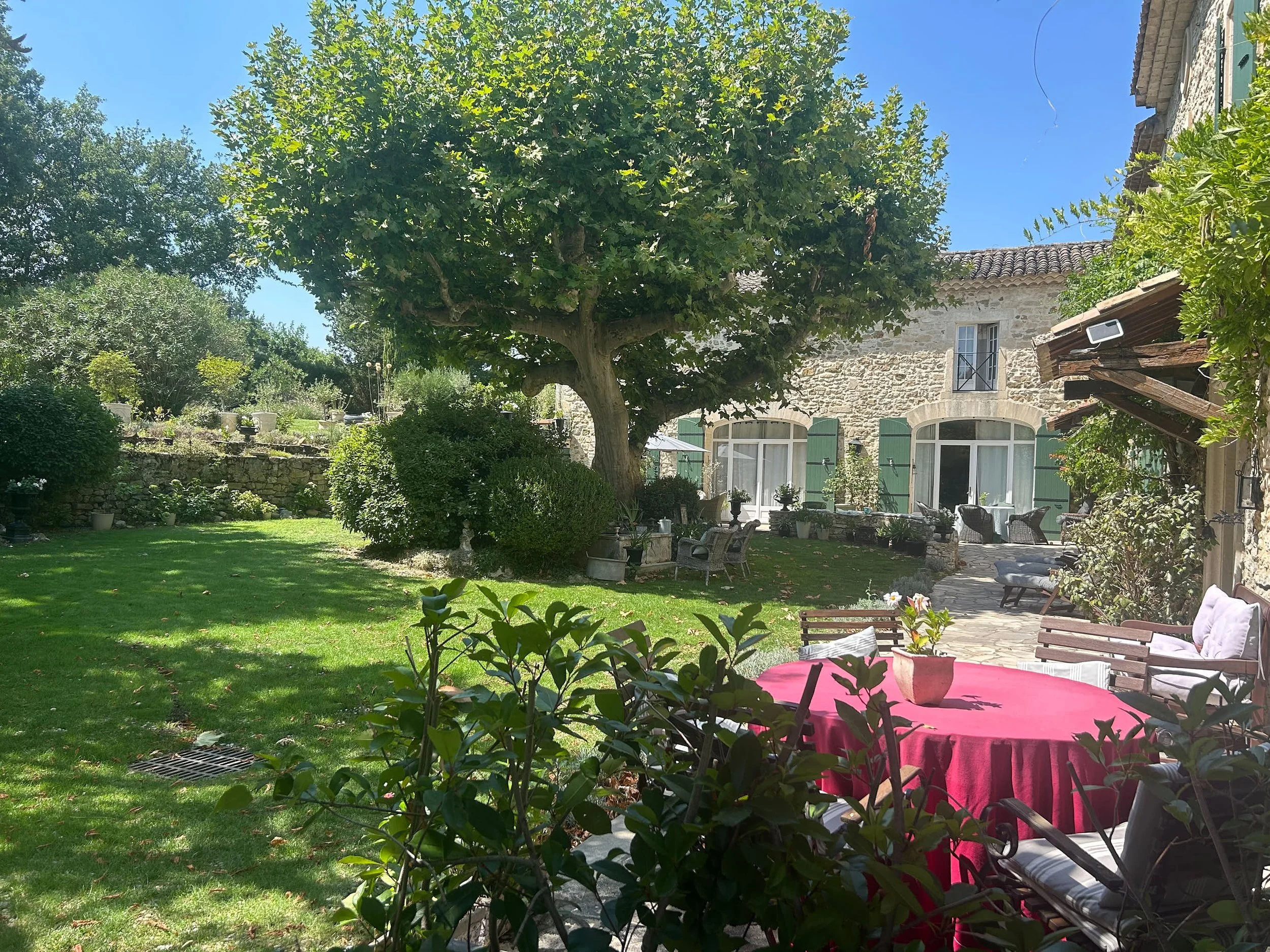 Garten mit Baum, Blumen, einer Terrasse mit Stühlen und einem runden Tisch mit roter Tischdecke, umgeben von Grün und einem alten Gebäude.