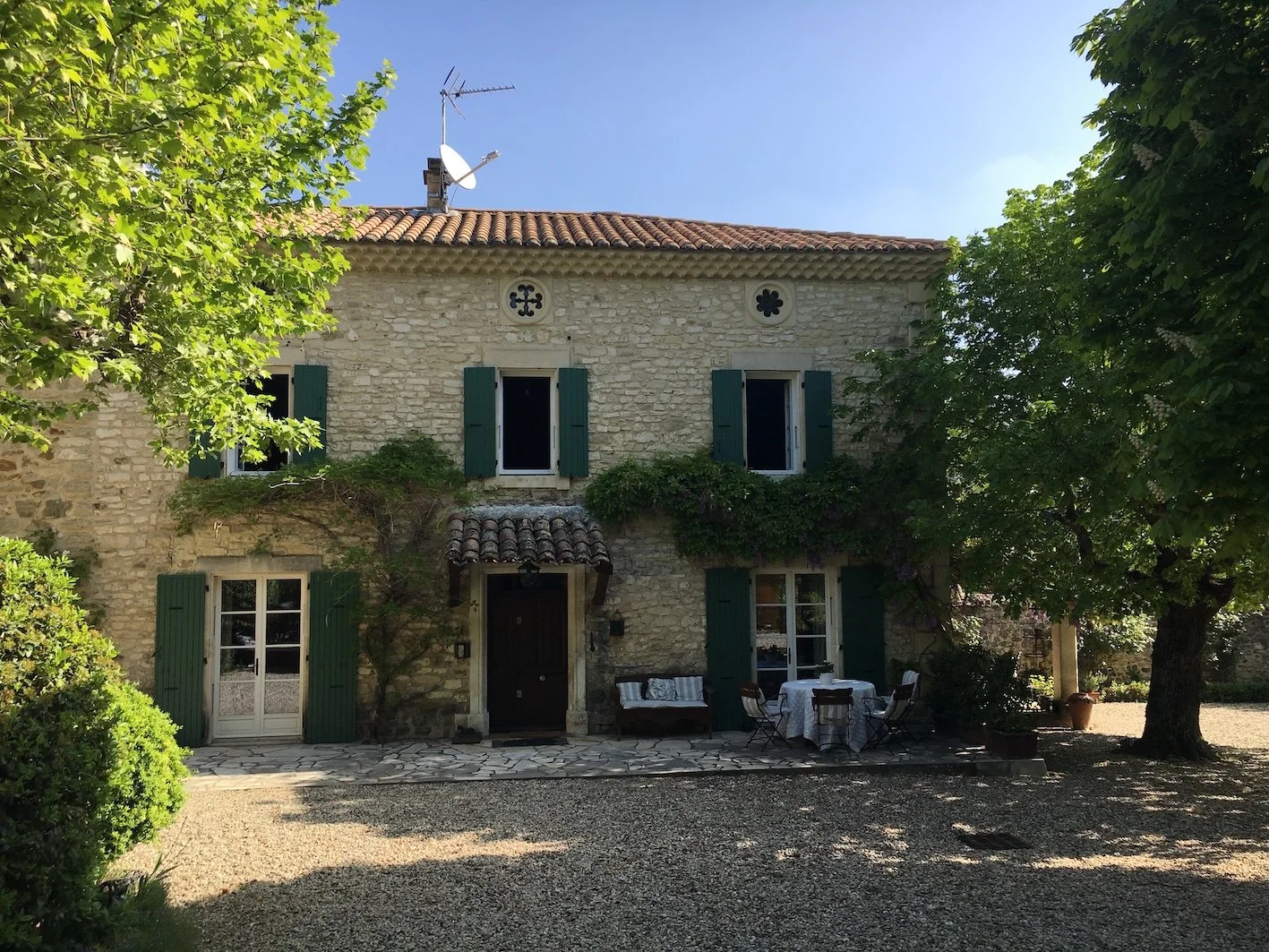 Ein altes zweistöckiges Steinhaus mit grünem Fensterläden, umgeben von Bäumen und einem Garten. Es gibt kleine Gartenmöbel vor dem Haus und eine Kiesfläche im Vordergrund.