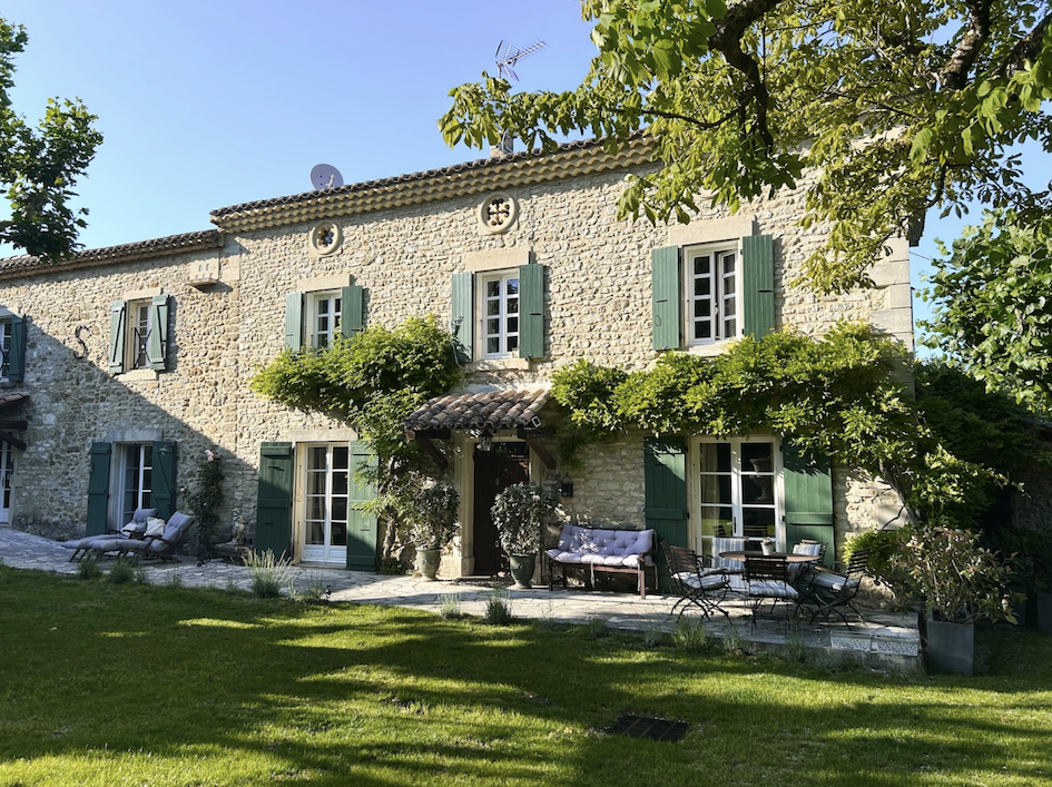 Ein zweistöckiges Haus mit Steinfassade, grünen Fensterläden und einer Terrasse mit Gartenmöbeln. Es gibt viel Grün mit Bäumen und Sträuchern im Garten.
