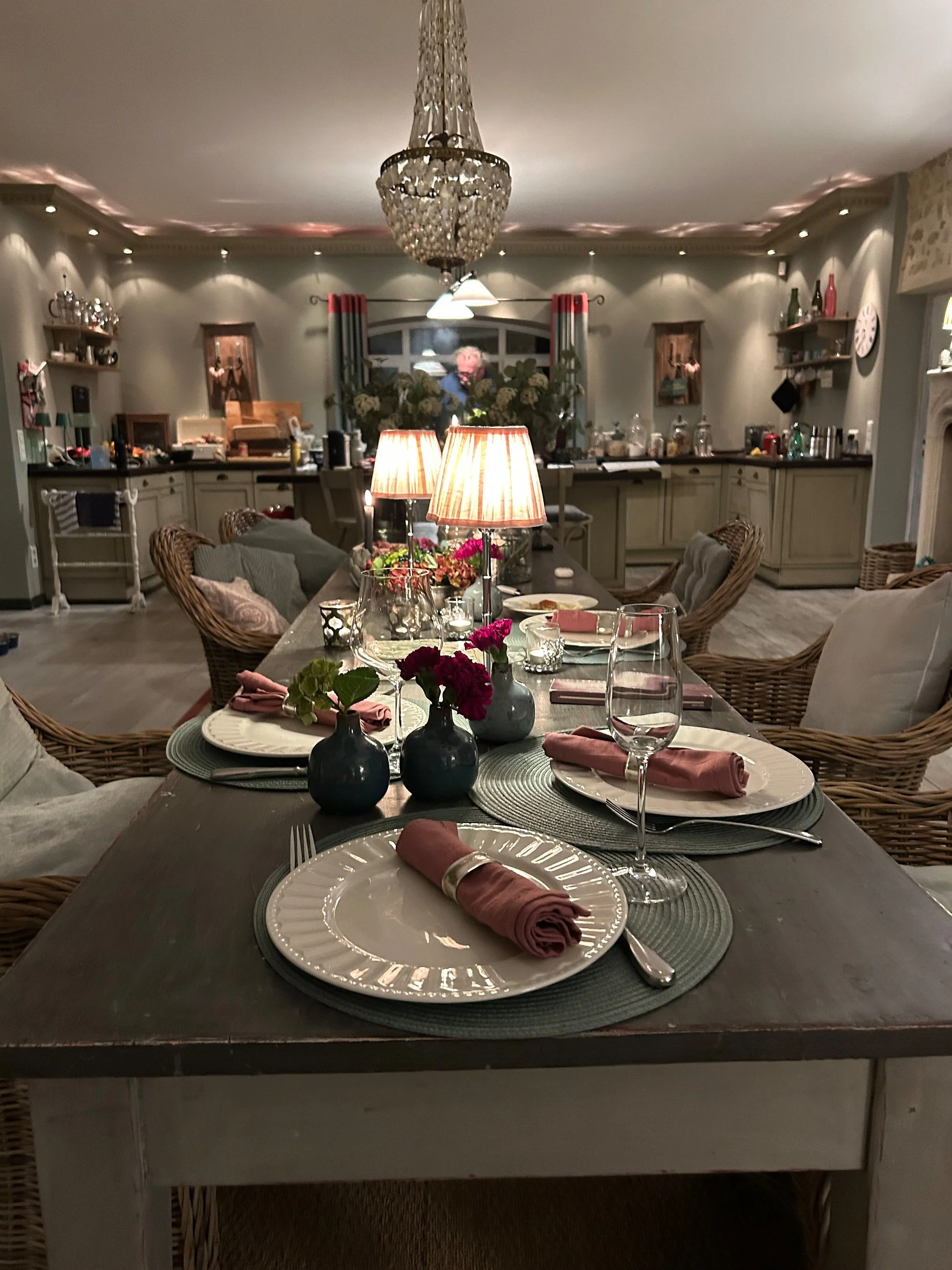 Ein festlich gedeckter Esstisch mit Tellern, Weingläsern, pinkfarbenen Servietten, Kerzen und Blumen, im Hintergrund eine Küche und ein Kronleuchter