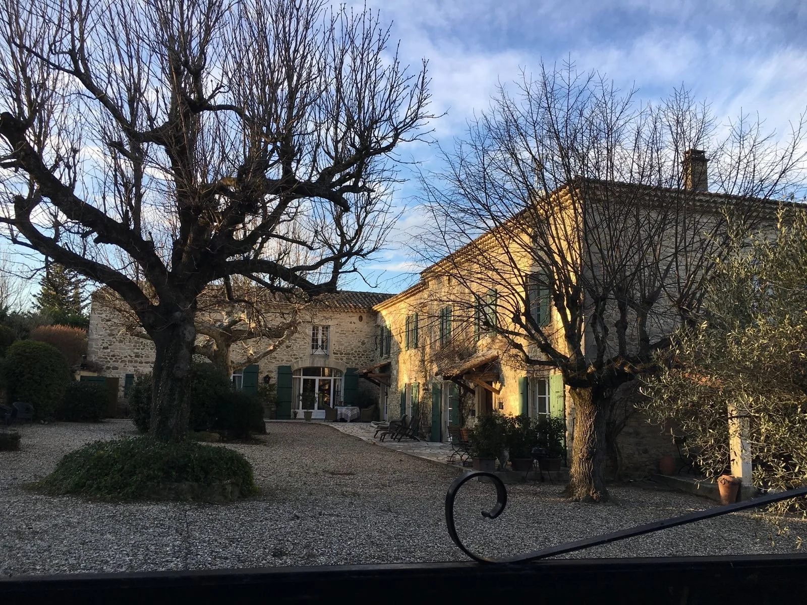 Historisches Haus mit Steinausführung und grünen Fensterläden, umgeben von Bäumen ohne Blätter, im Sonnenlicht bei klarem Himmel.