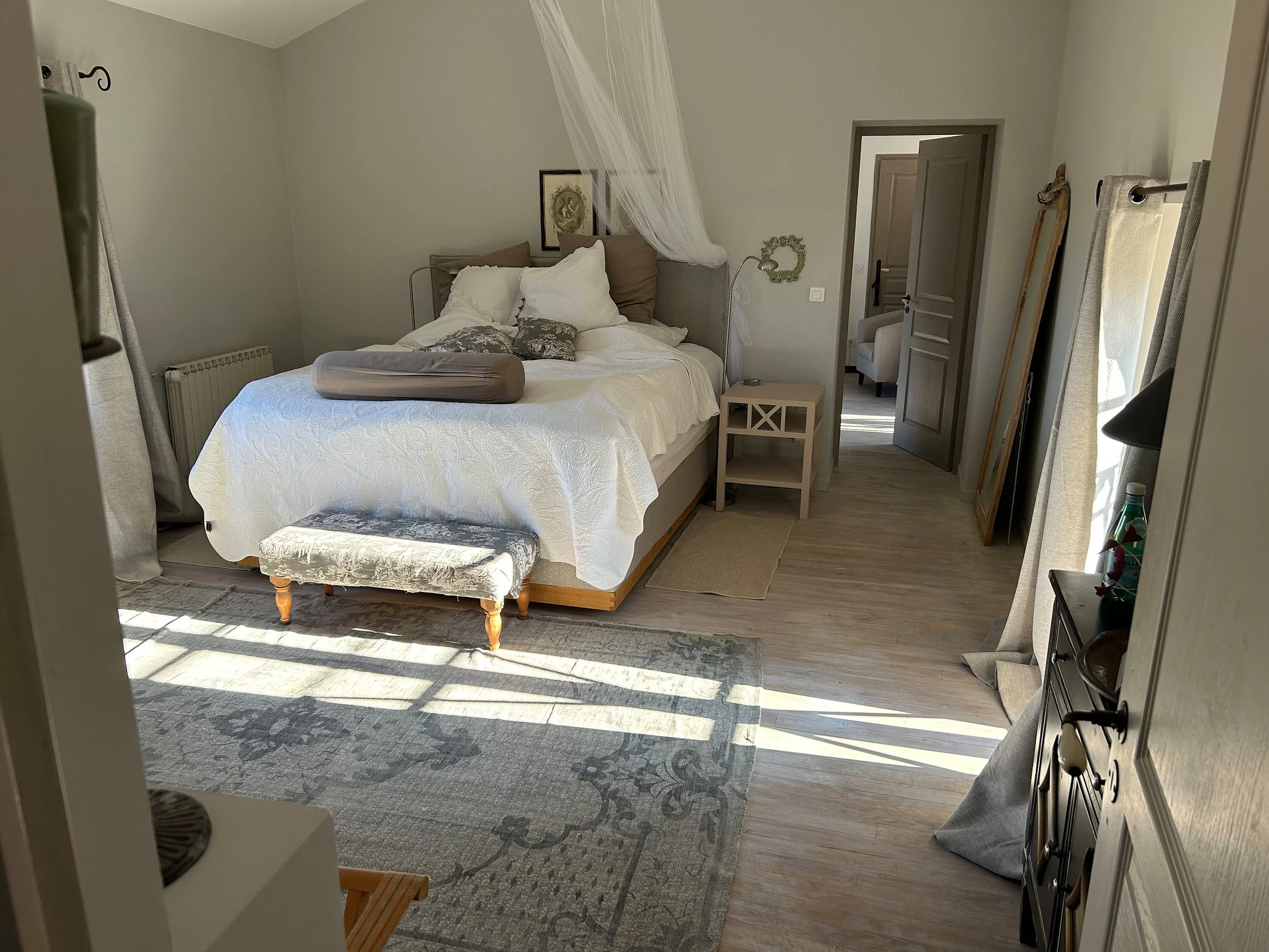 Ein gemütliches Schlafzimmer mit einem großen Bett, weißen Bettwäsche und mehreren Kissen. Es gibt einen kleinen Hocker am Fußende des Bettes, einen Nachttisch neben dem Bett und ein Sofa im Hintergrund. Das Zimmer ist hell erleuchtet durch Sonnenlic