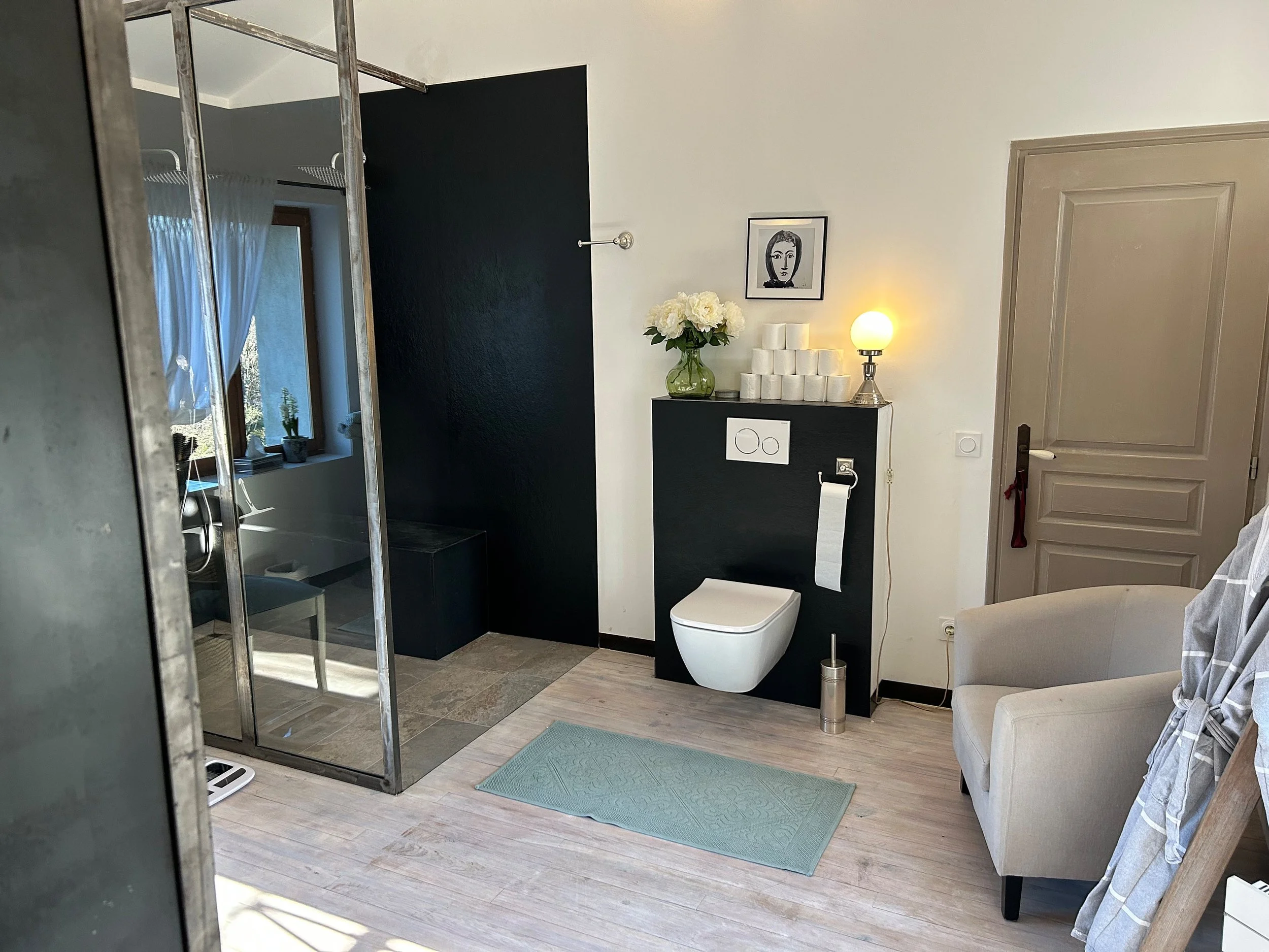 Ein modern eingerichtetes Badezimmer mit begehbarer Dusche, einem Toilettenbereich mit schwarzen Wandpaneelen, einem hellen Sessel, einem Schrank mit Blumen und Kerzen, einem Wandbild und großen Fenstern mit weißen Vorhängen.