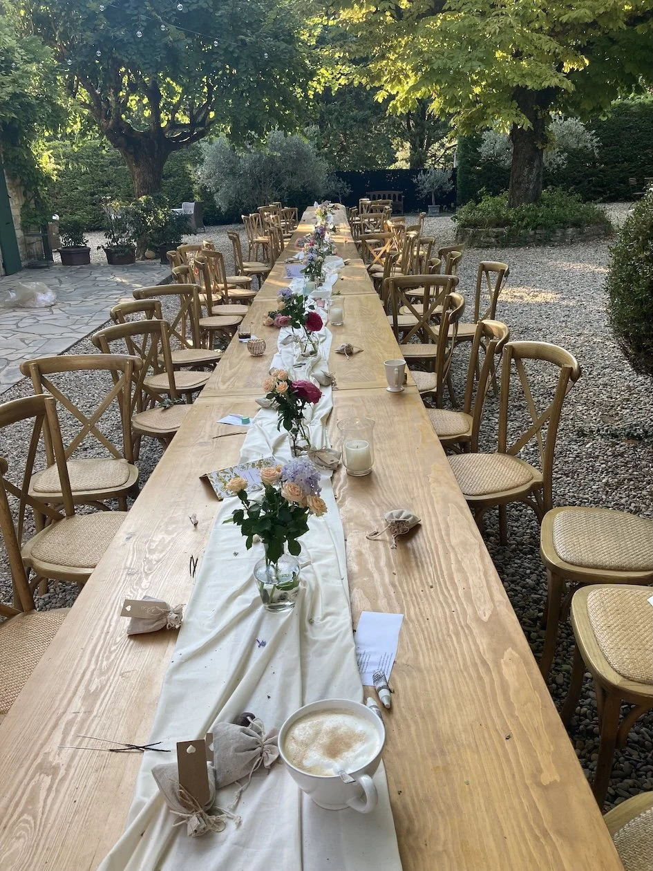 Langer Holztisch mit Blumen, Kerzen und Platzdeckchen im Garten, umgeben von Stühlen, für eine Veranstaltung oder Feier. Im Hintergrund Bäume und Pflanzen.