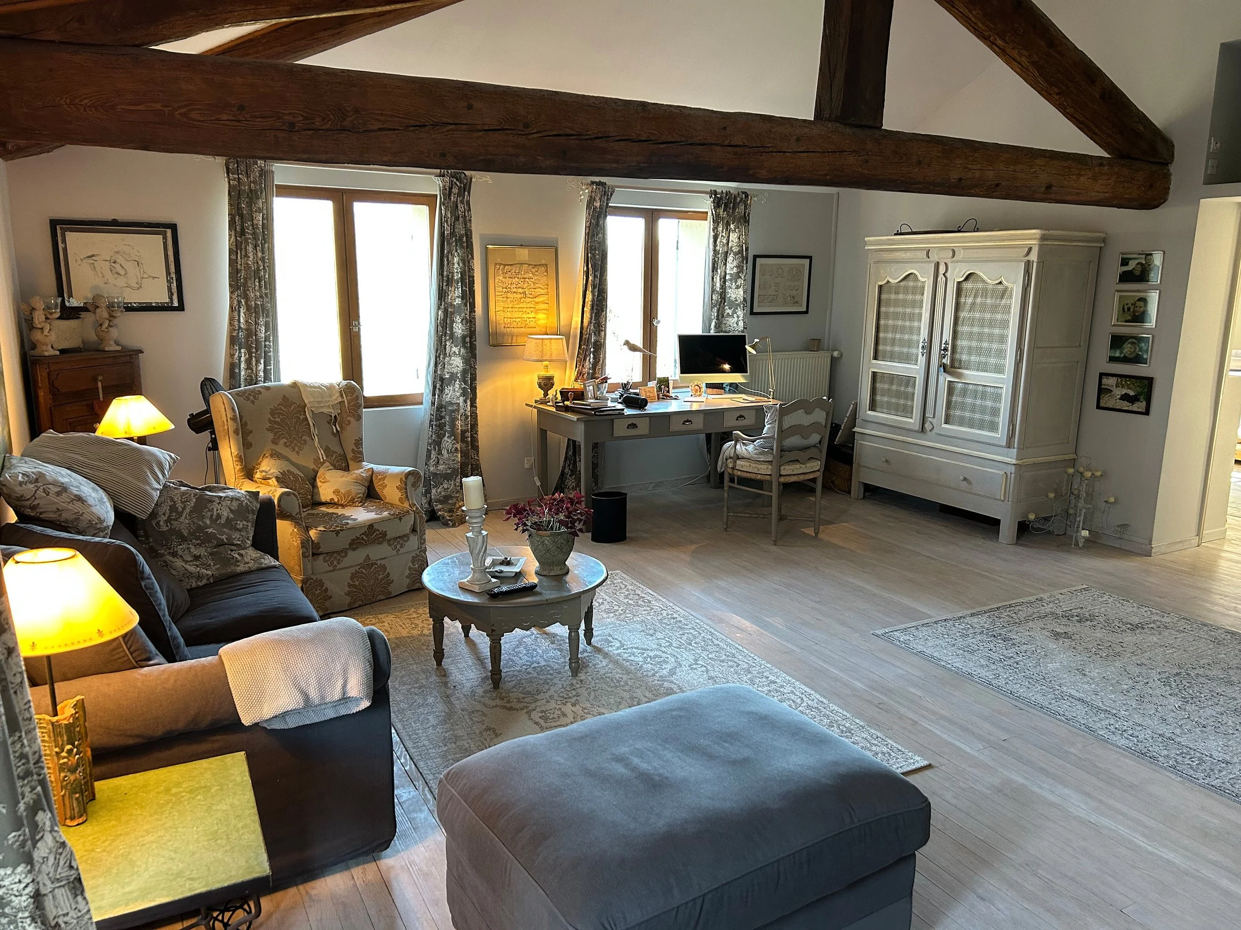 Gemütliches Wohnzimmer mit braunem Sofa, weißen Sessel, Couchtisch mit Blumen und Kerzen, Schreibtisch mit Computer, weißer Kommode, Holzdecke und mehreren Fenstern mit Vorhängen.