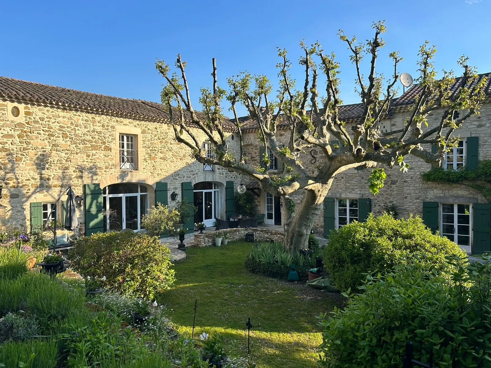 Ein alter Stein-Häuser mit grünen Fensterläden, umgeben von einem Garten mit Bäumen und Sträuchern, bei Sonnenlicht und klarem Himmel.