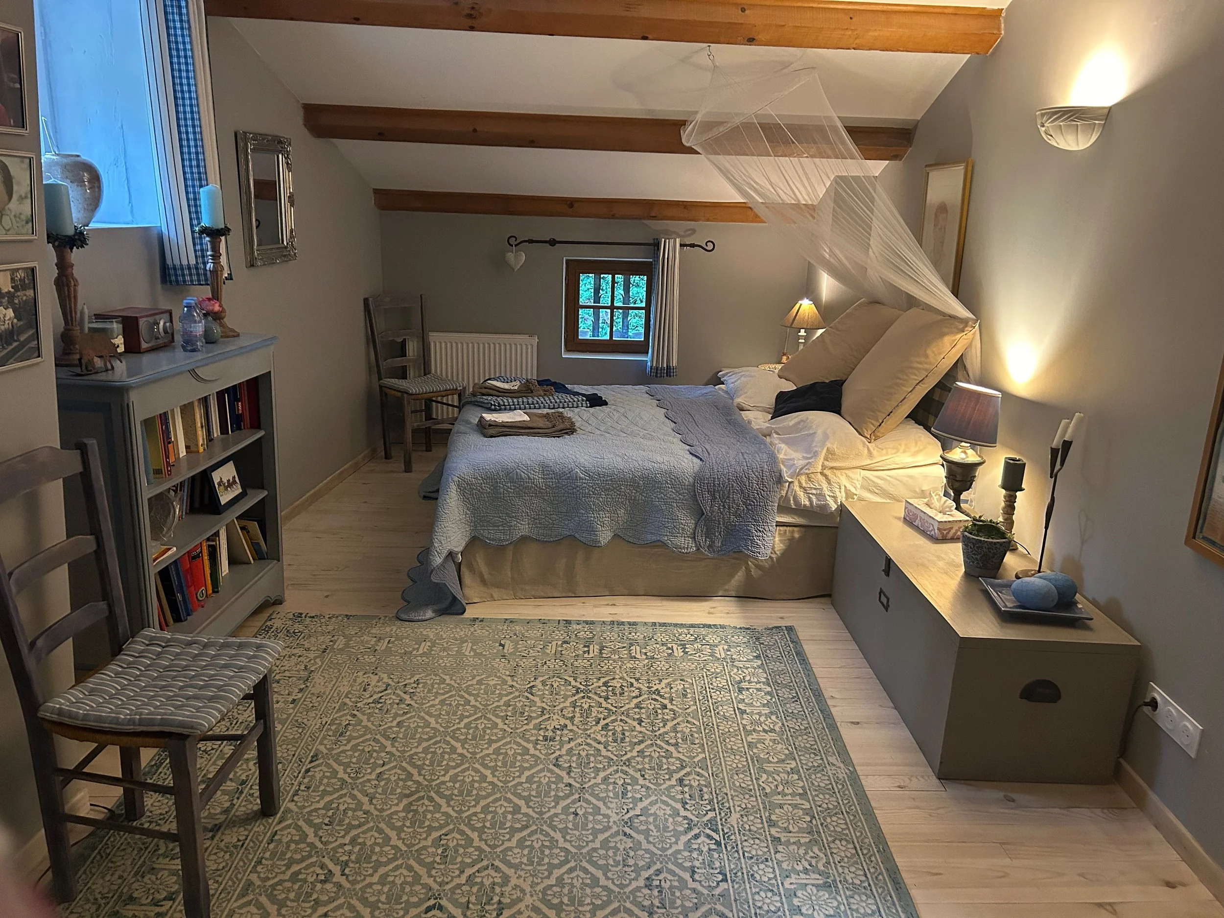 Gemütliches Schlafzimmer mit Doppelbett, Bettwäsche, Nachttischen mit Lampen, Bücherregal, Holzdecke, Teppich, kleine Fenster, und Dekorationen.