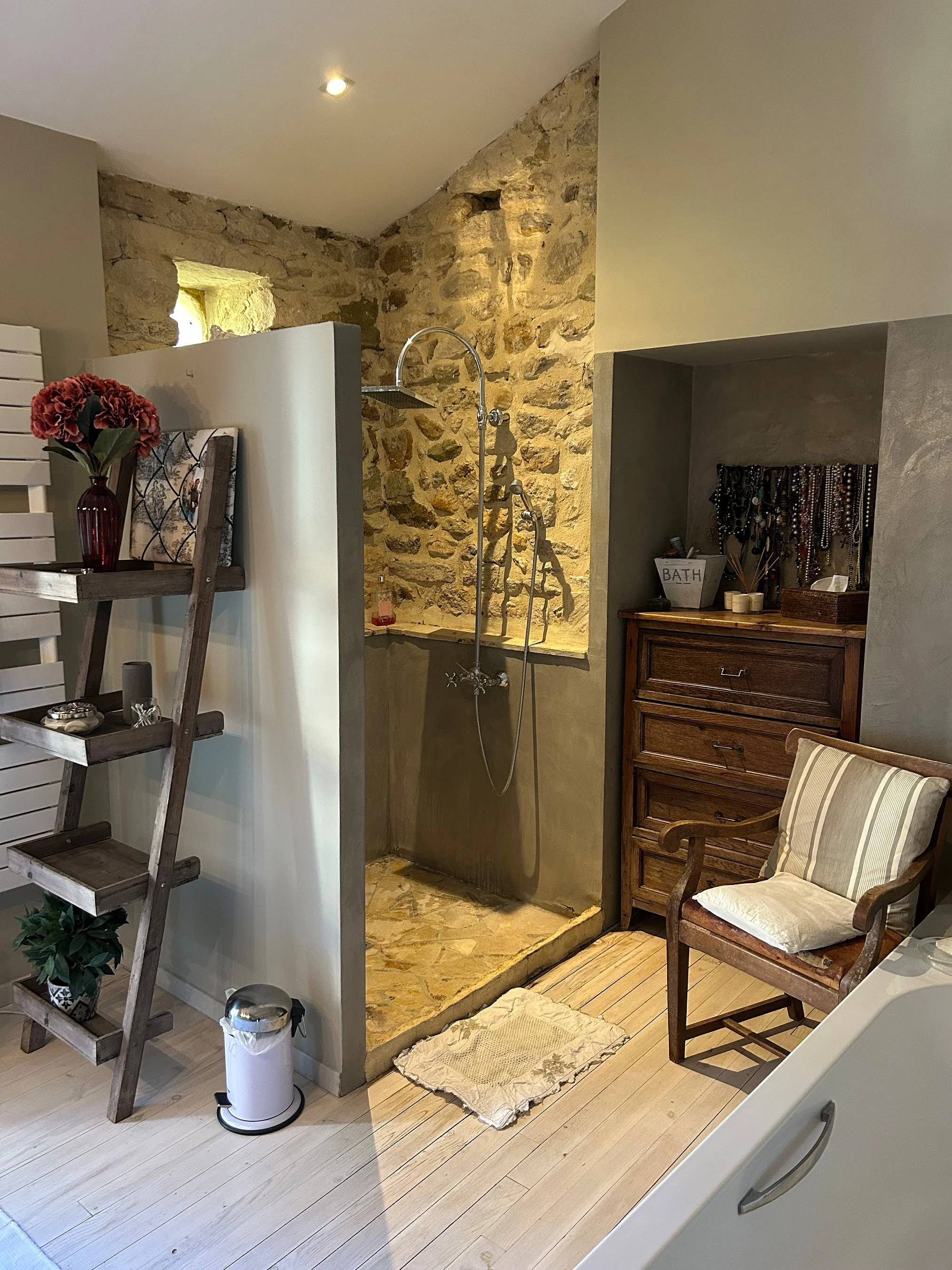 Badezimmer mit offener Walk-In-Dusche, rustikaler Steinwand und Holzfußboden, mit antiker Holzkommode, Sessel mit Kissen, Pflanzen und dekorativen Gegenständen.