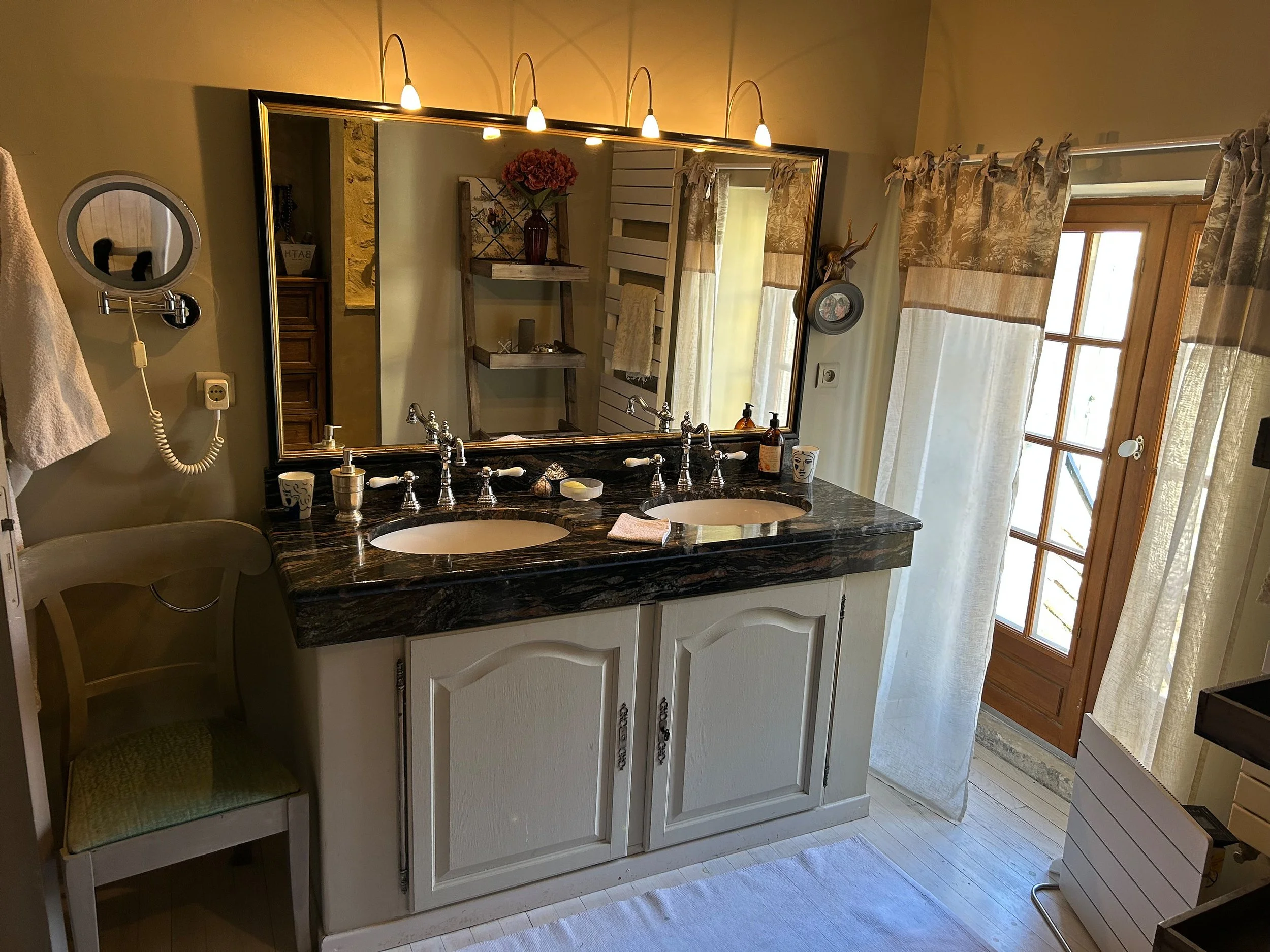 Ein Badezimmer mit Doppel-Waschtisch, dunklem Marmor, großem Spiegel und Fenster mit Vorhängen