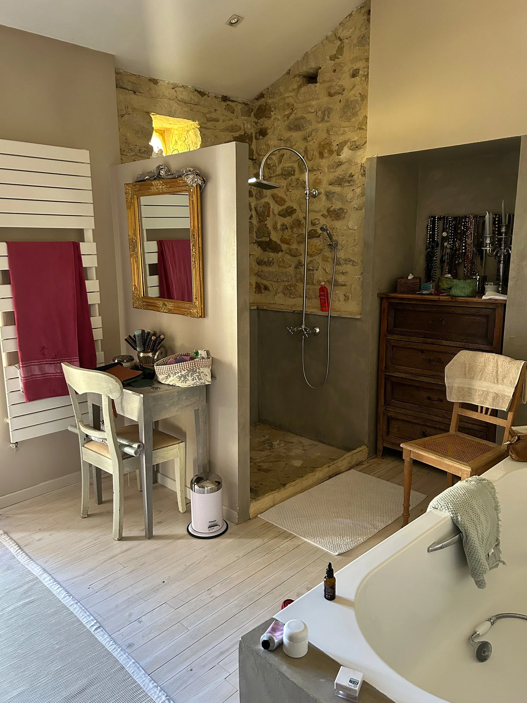 Ein Badezimmer mit offener Dusche aus Stein, einem Waschtisch, einem Spiegel, einem Stuhl, einem Badebereich mit Badewanne und verschiedenen Pflegeartikeln.