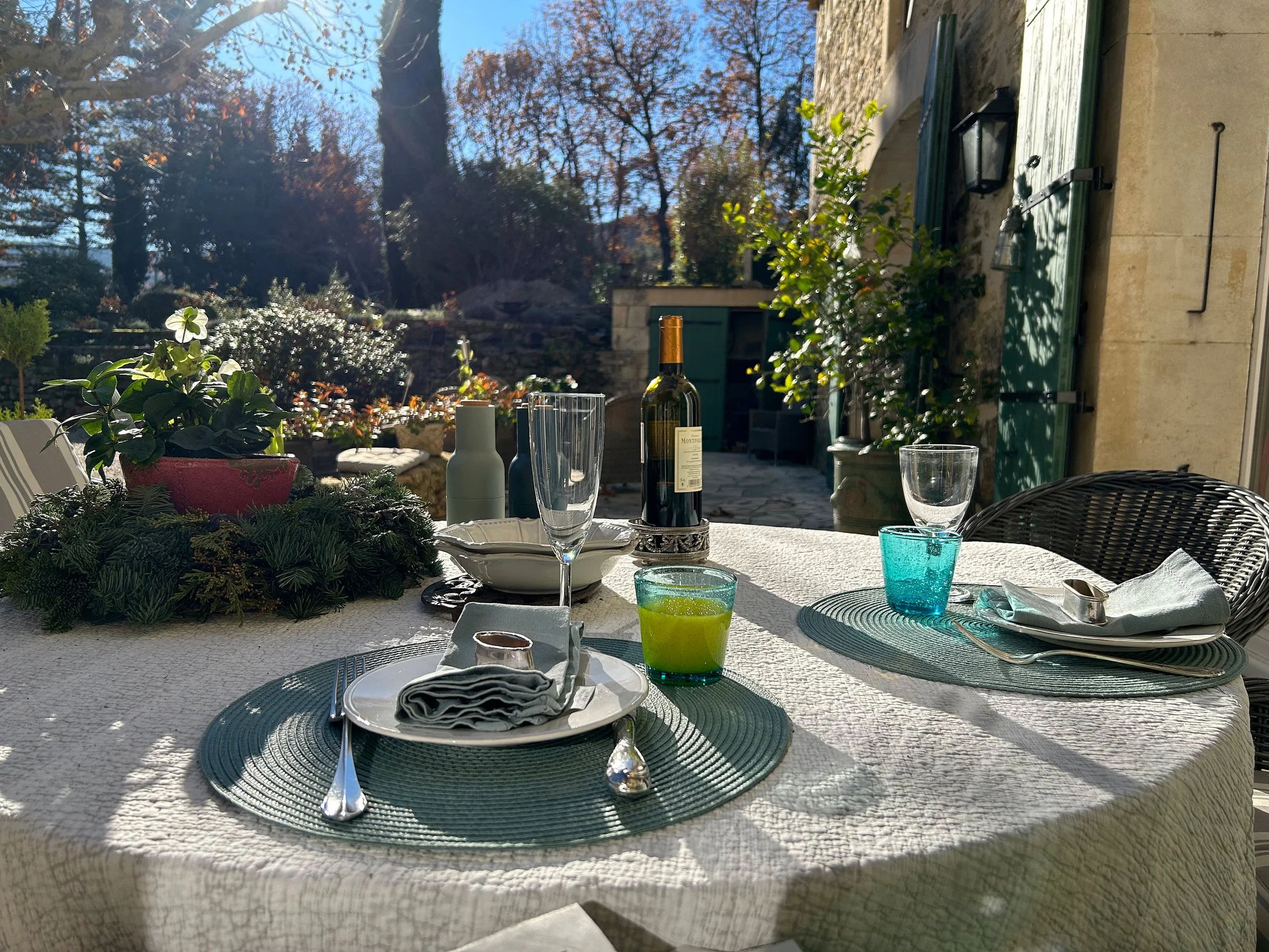 Ein gedeckter Esstisch im Freien mit Geschirr, Gläsern, Besteck, einer Weinflasche und einer grünen Kerze, umgeben von Pflanzen und Bäumen bei Sonnenlicht.