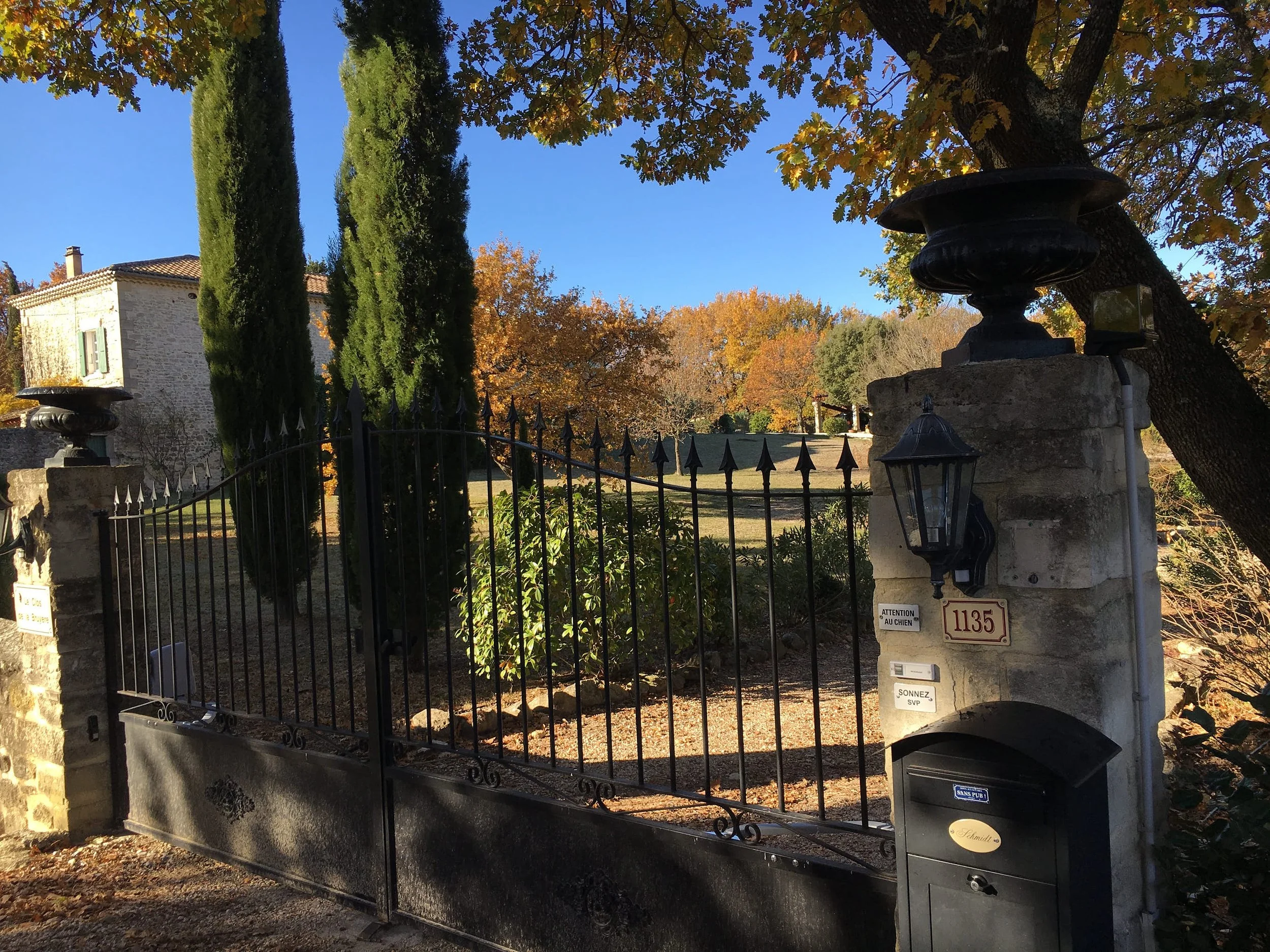 Ein schmiedeeisernes Tor vor einem Haus mit einem Zaun und bestimmten Hausnummern, umgeben von Bäumen mit Herbstblättern.
