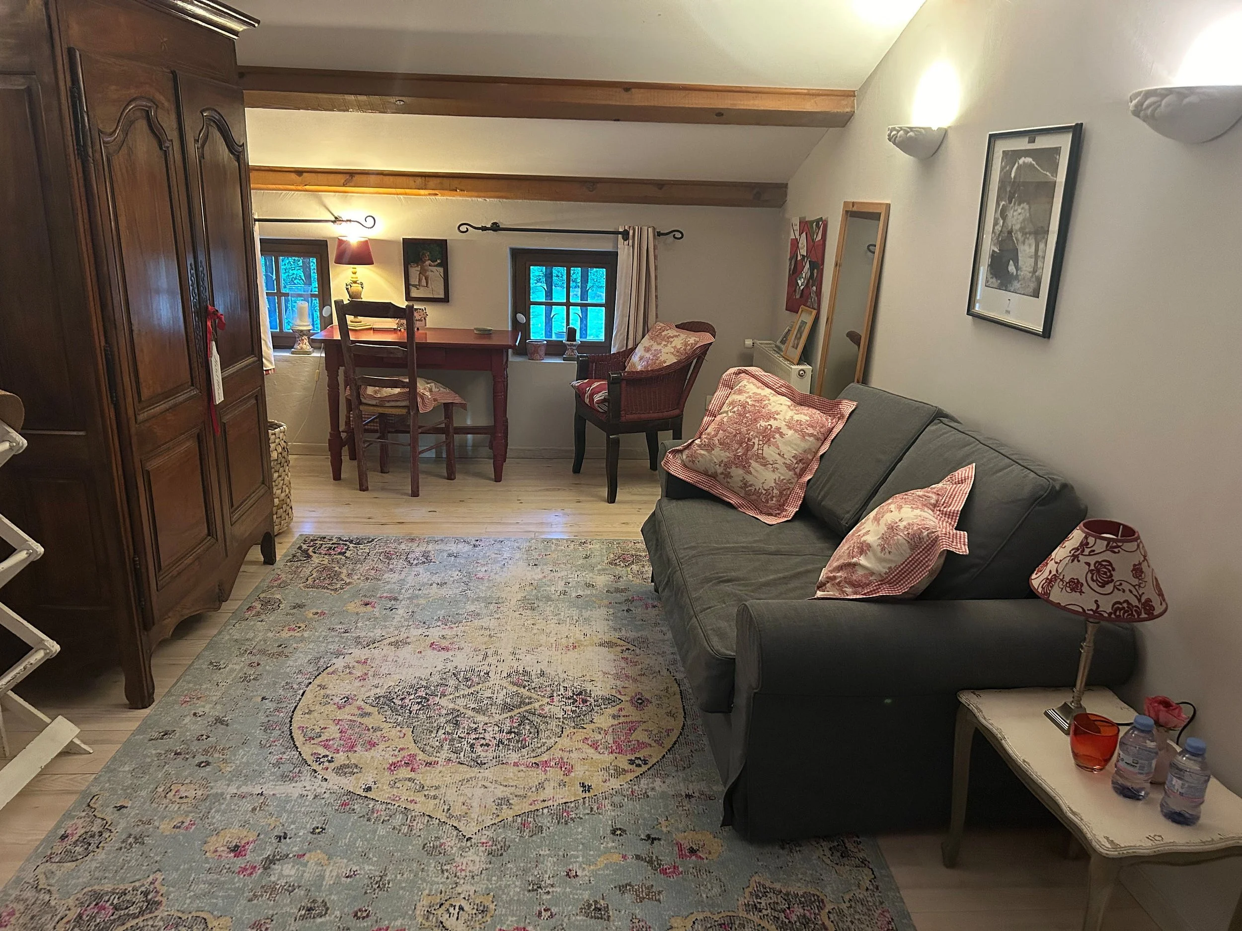 Ein gemütliches Wohnzimmer mit dunklem Sofa, mehreren Kissen, Teppich, Holzschrank, kleinen Tischen, Sesseln, Bildern an der Wand, zwei kleinen Fenstern, Lichter und Dekorationen, insgesamt warm beleuchtet.