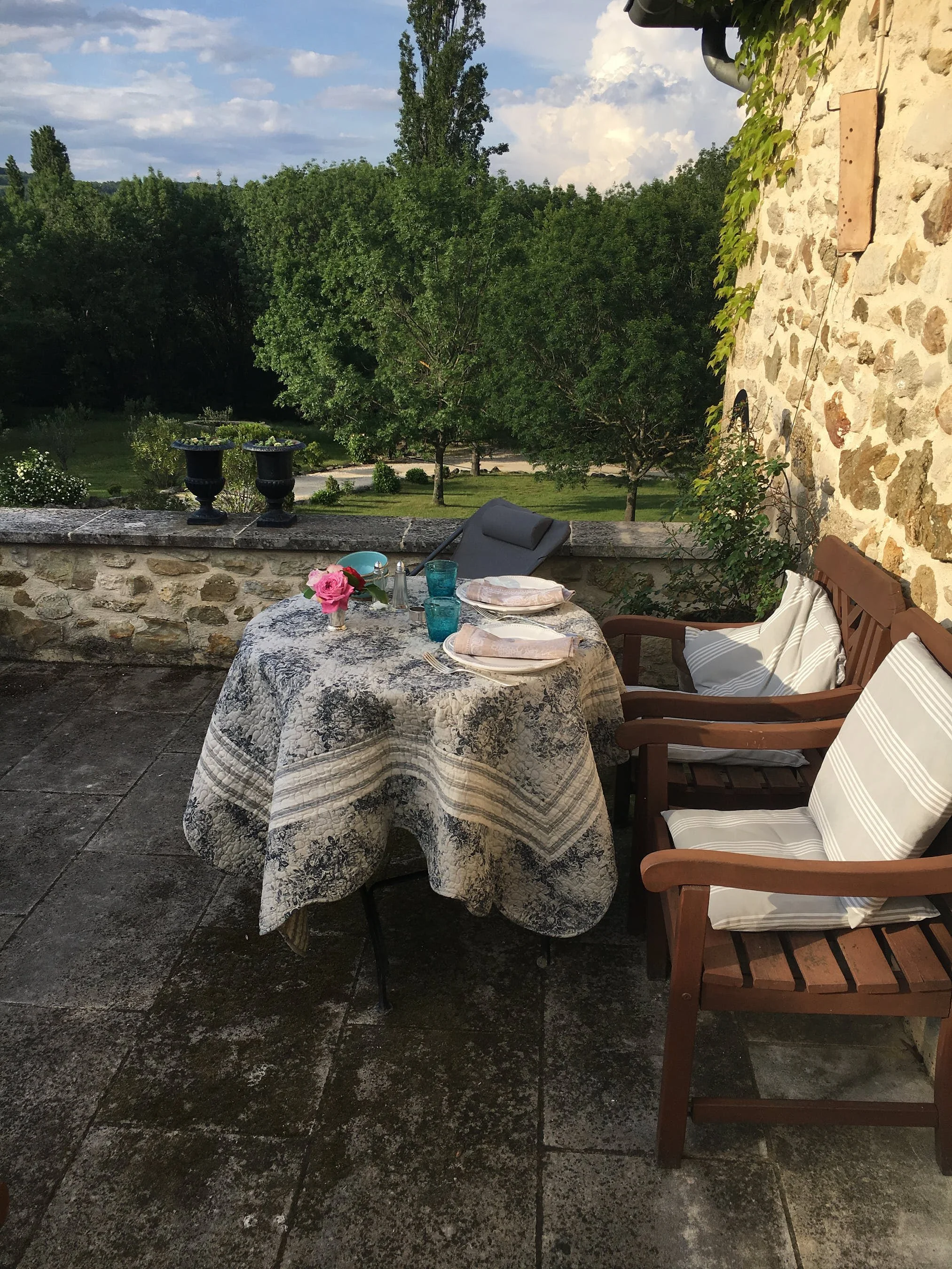 Ein Tisch mit einem Blumenstrauß, gedeckt für ein Essen, auf einem terrassierten Balkon mit Blick auf einen Garten voller Bäume, während draußen die Sonne scheint.